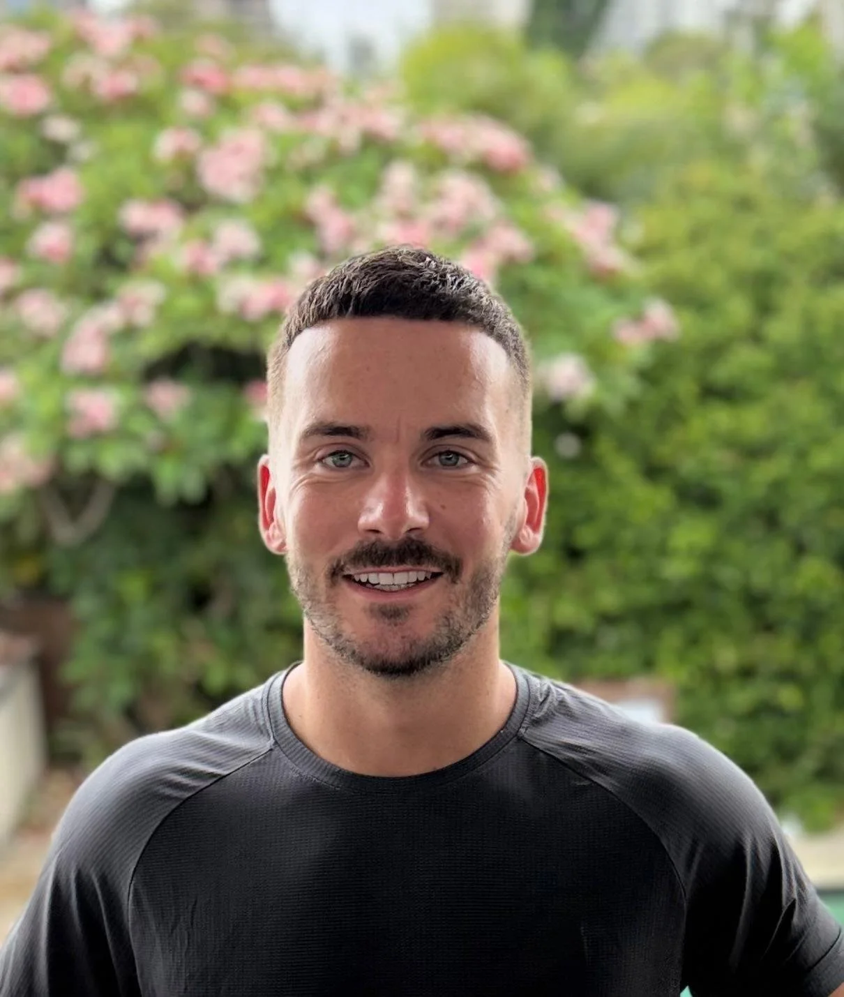 A smiling man with short dark hair and a beard, wearing a black athletic shirt, standing outdoors with green trees and pink flowers in the background.