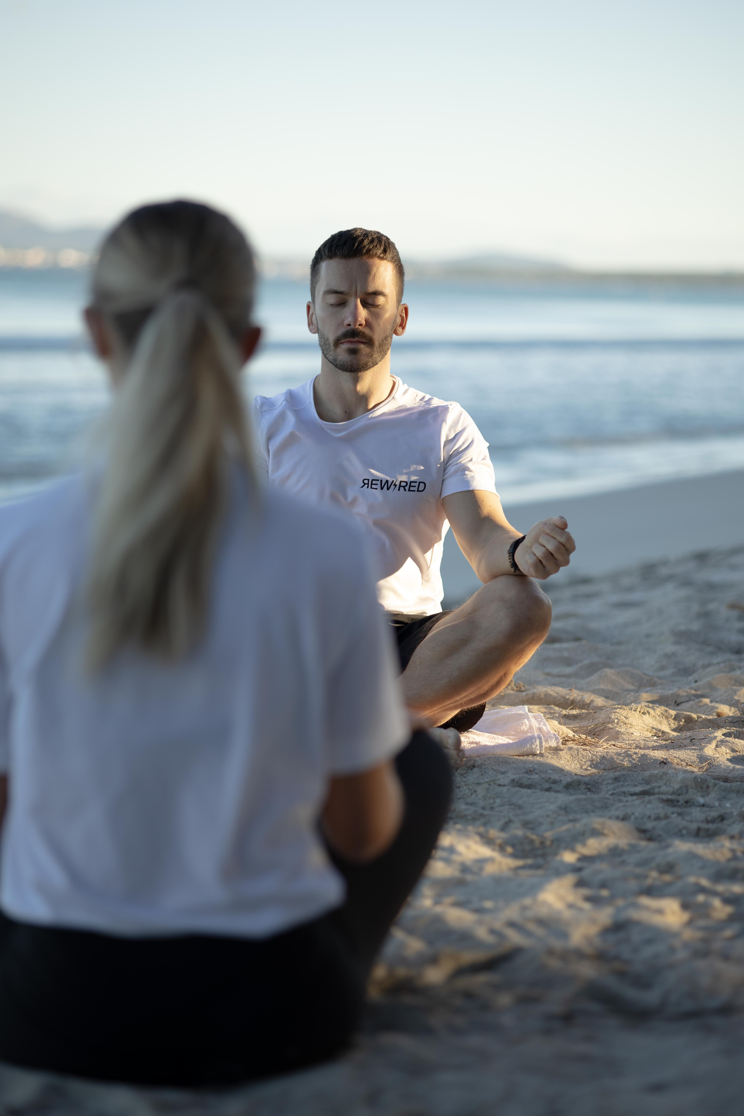 A man and a woman meditating on a beach at sunset, sitting cross-legged on the sand with the ocean and mountains in the background.