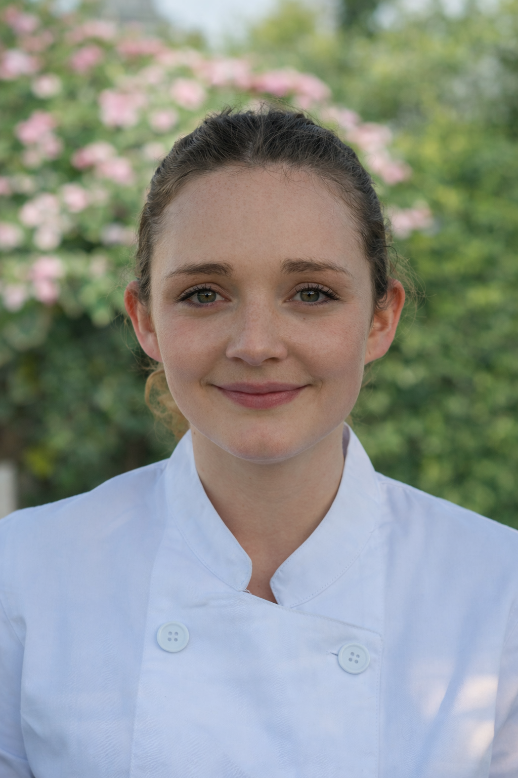 Young woman in a white chef's coat outdoors with greenery and pink flowers in the background