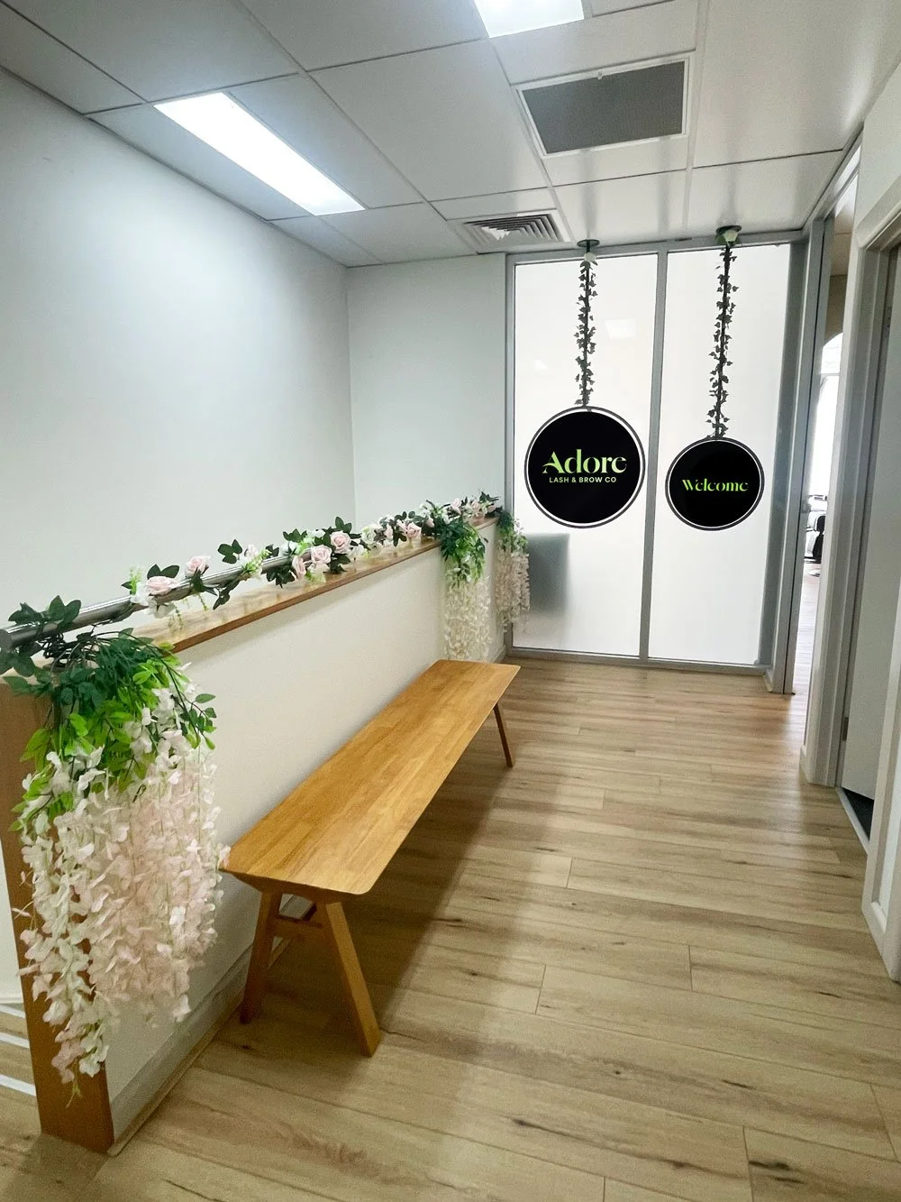 Empty waiting area with a wooden bench, decorated with pink and white flowers and greenery along the wall. Two circular signs hang from the ceiling, one reading 'Adore Lash & Brow Co' and the other 'Welcome,' in front of a glass partition with frosted upper sections.