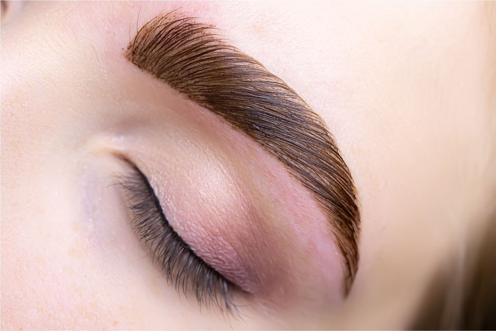 Close-up of a person's closed eye showing well-groomed, laminated eyebrows, long eyelashes, and light eye makeup.