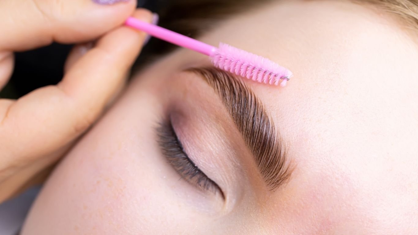 Close-up of a person's face with closed eyes as someone cleans or shapes their eyebrow with a pink brush.