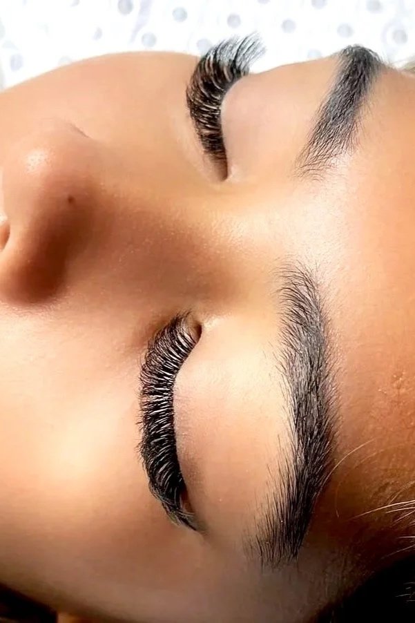 Close-up of a woman's face with eyes closed, showing long, thick eyelashes, neatly groomed eyebrows, and smooth skin.