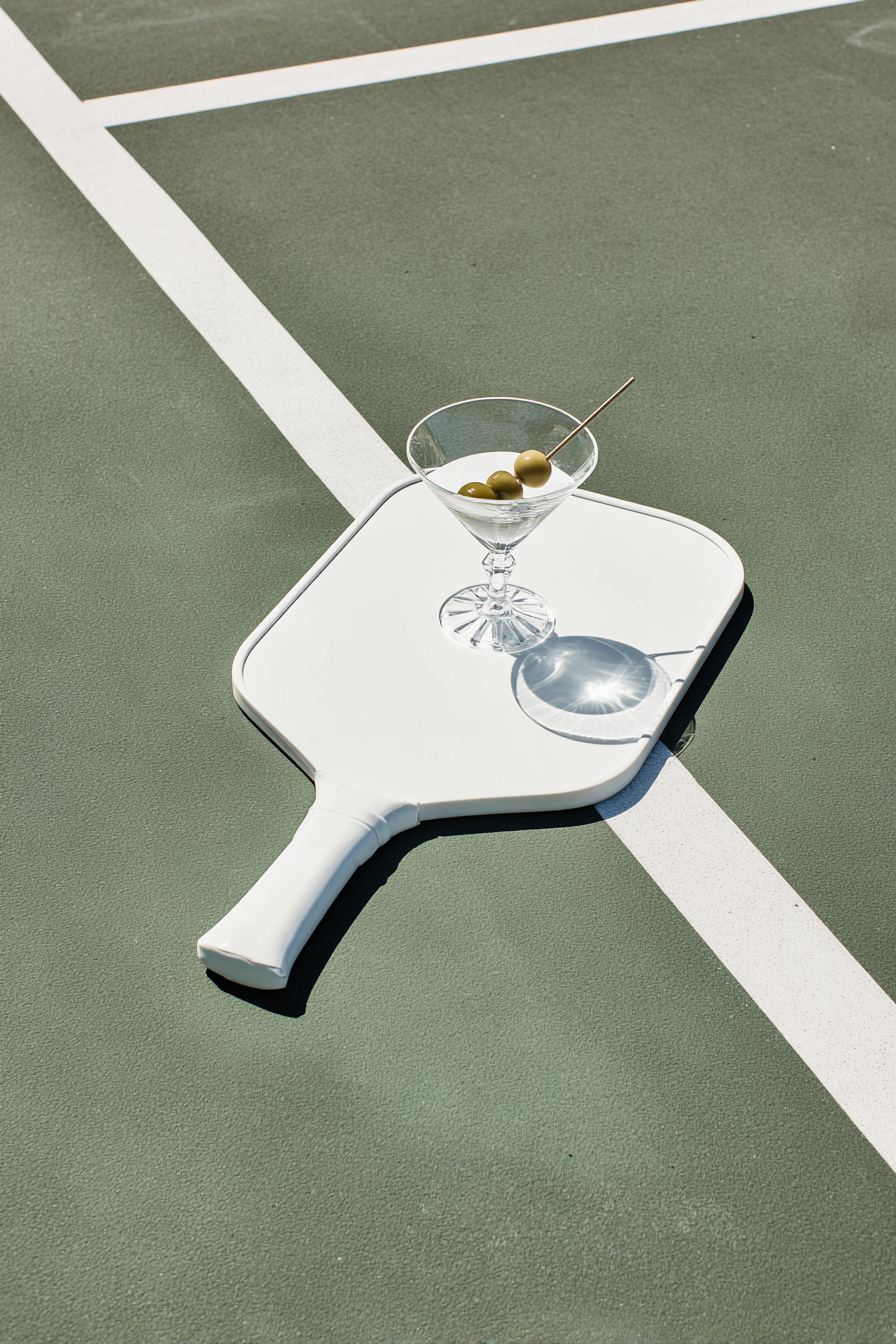 A ping pong paddle with a martini glass containing green olives on a court surface.