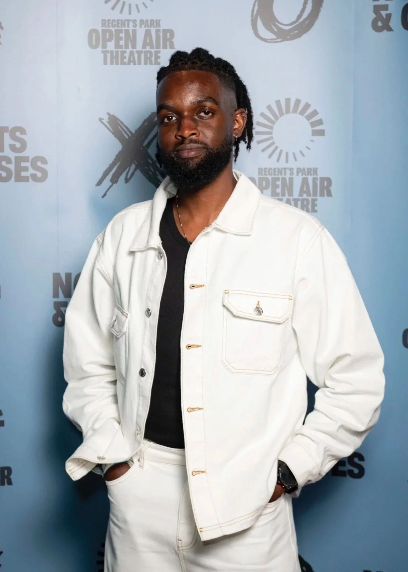 A black man in his 30s with locs wearing a baggy white denim co-ord with a black vest posing for a photo in front of the Noughts & Crosses blue backdrop at Regent’s Park Open Air Theatre