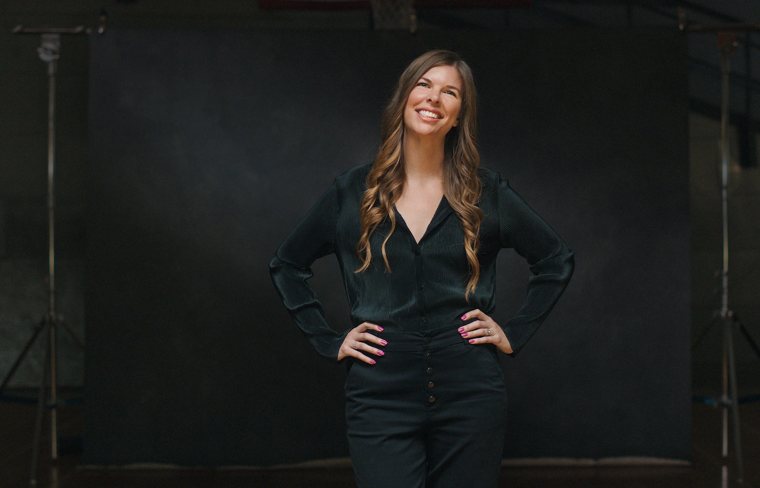 A woman with long, wavy brown hair, wearing a black shirt and black pants, smiling confidently with hands on her hips in a studio setting.