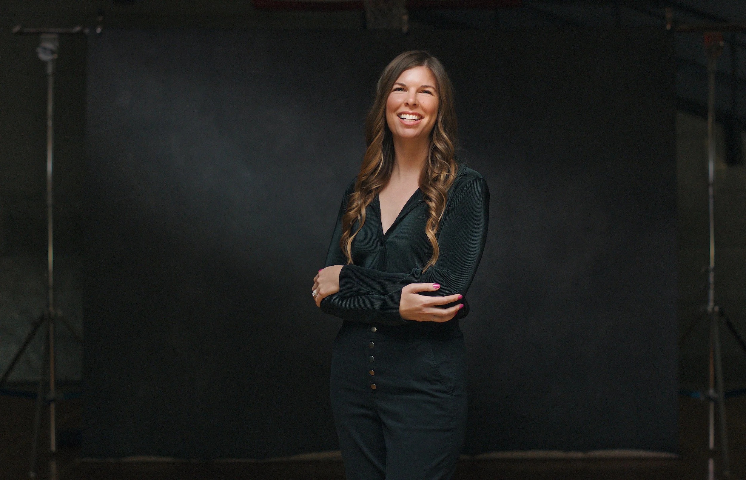 A young woman with long wavy brown hair smiling and standing with her arms crossed in front of a dark backdrop in a studio setting.