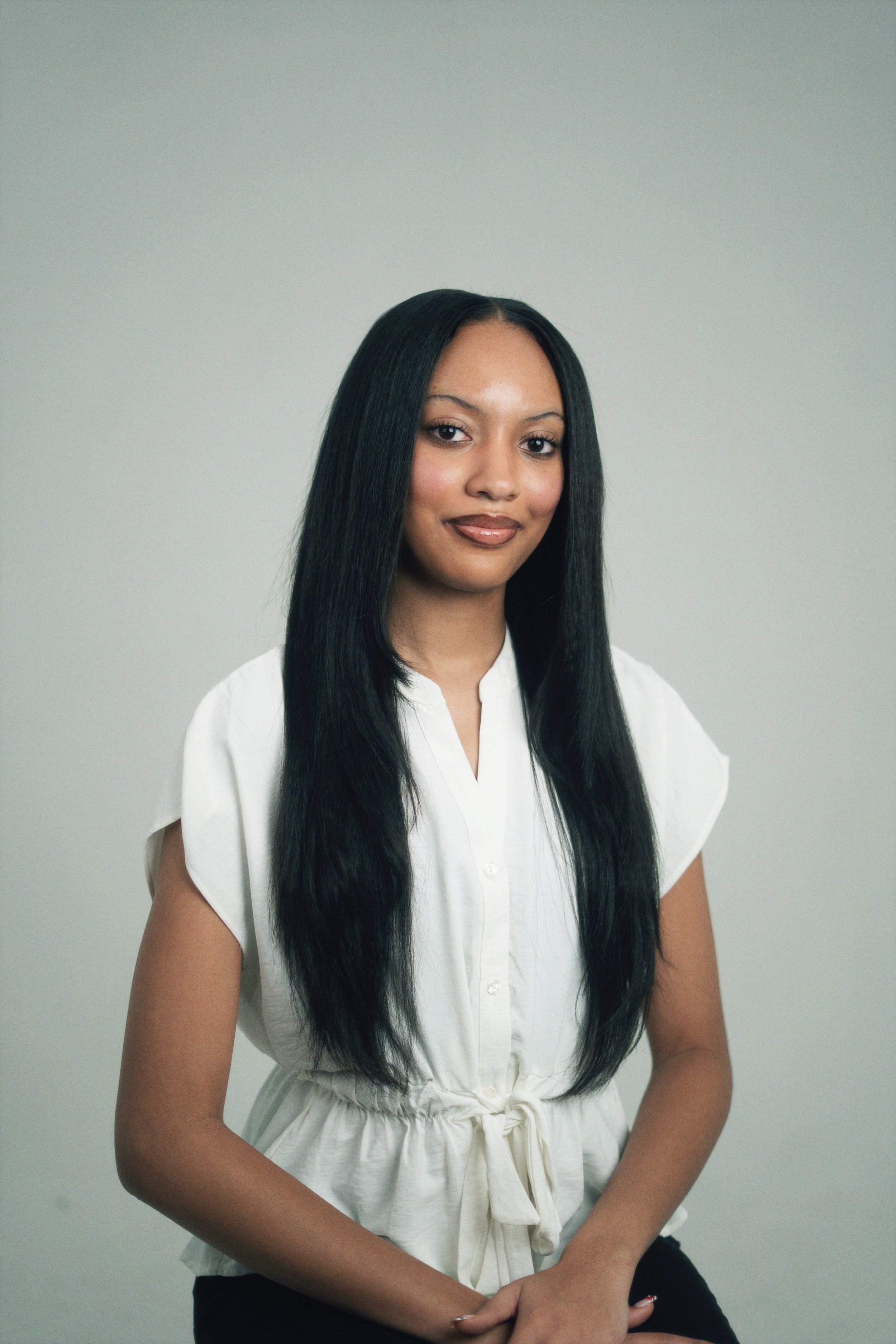 A young woman with long black hair, wearing a white blouse and smiling slightly, standing against a plain light grey background.