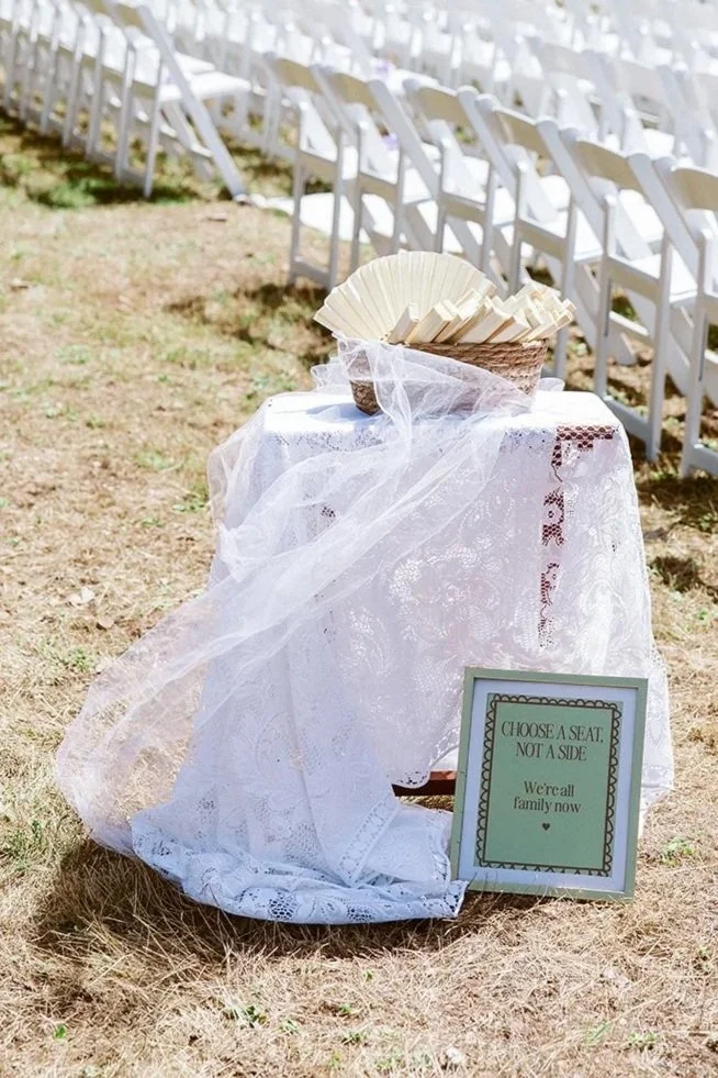 Outdoor wedding setup with rows of white chairs facing a green natural backdrop, a small table covered with white lace holding a basket of paper fans, and a sign that reads, 'Choose a seat, not a side. We're all family now.'