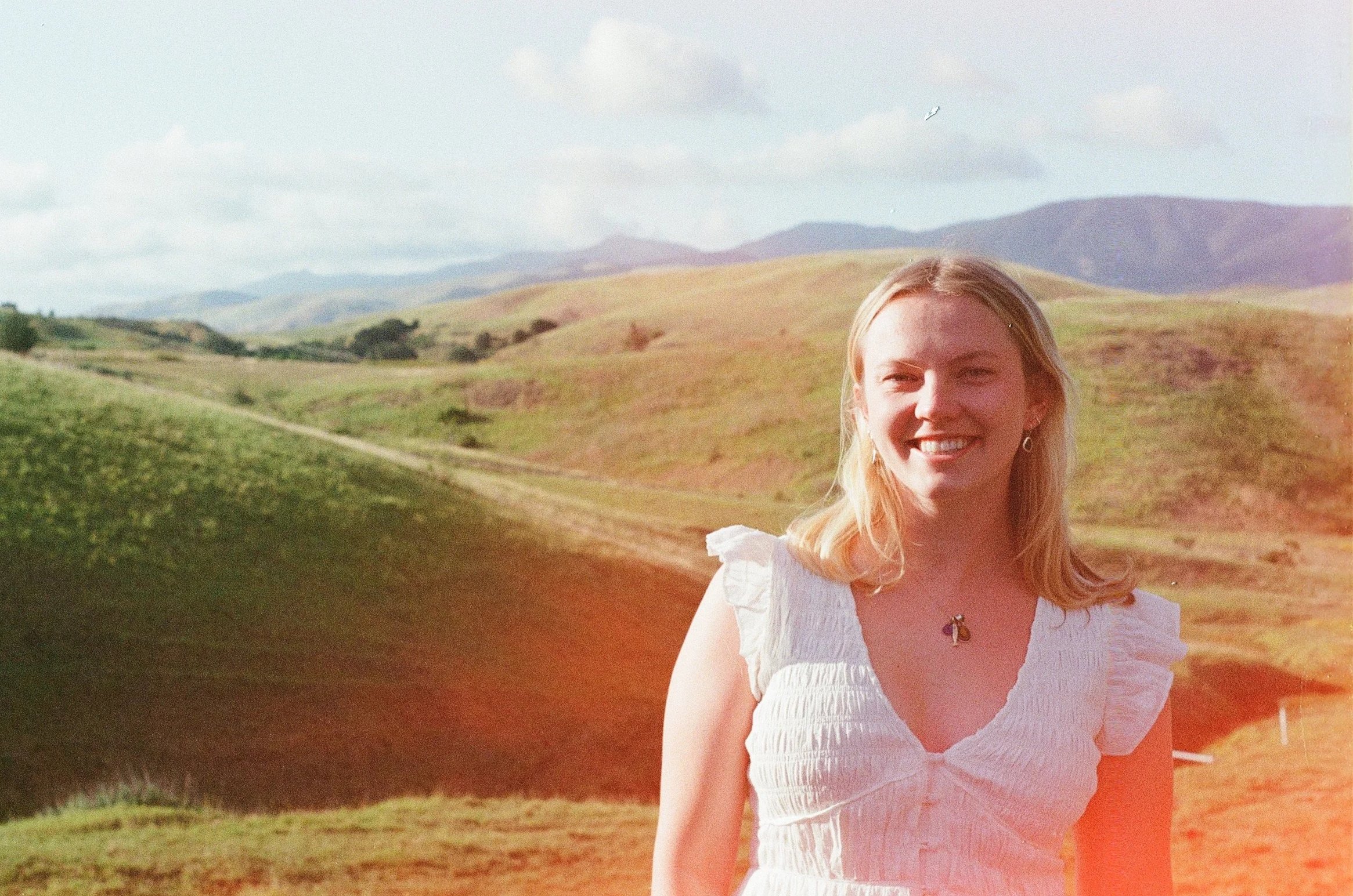 A young woman with blond hair smiling outdoors in a scenic landscape of rolling hills and mountains under a partly cloudy sky.