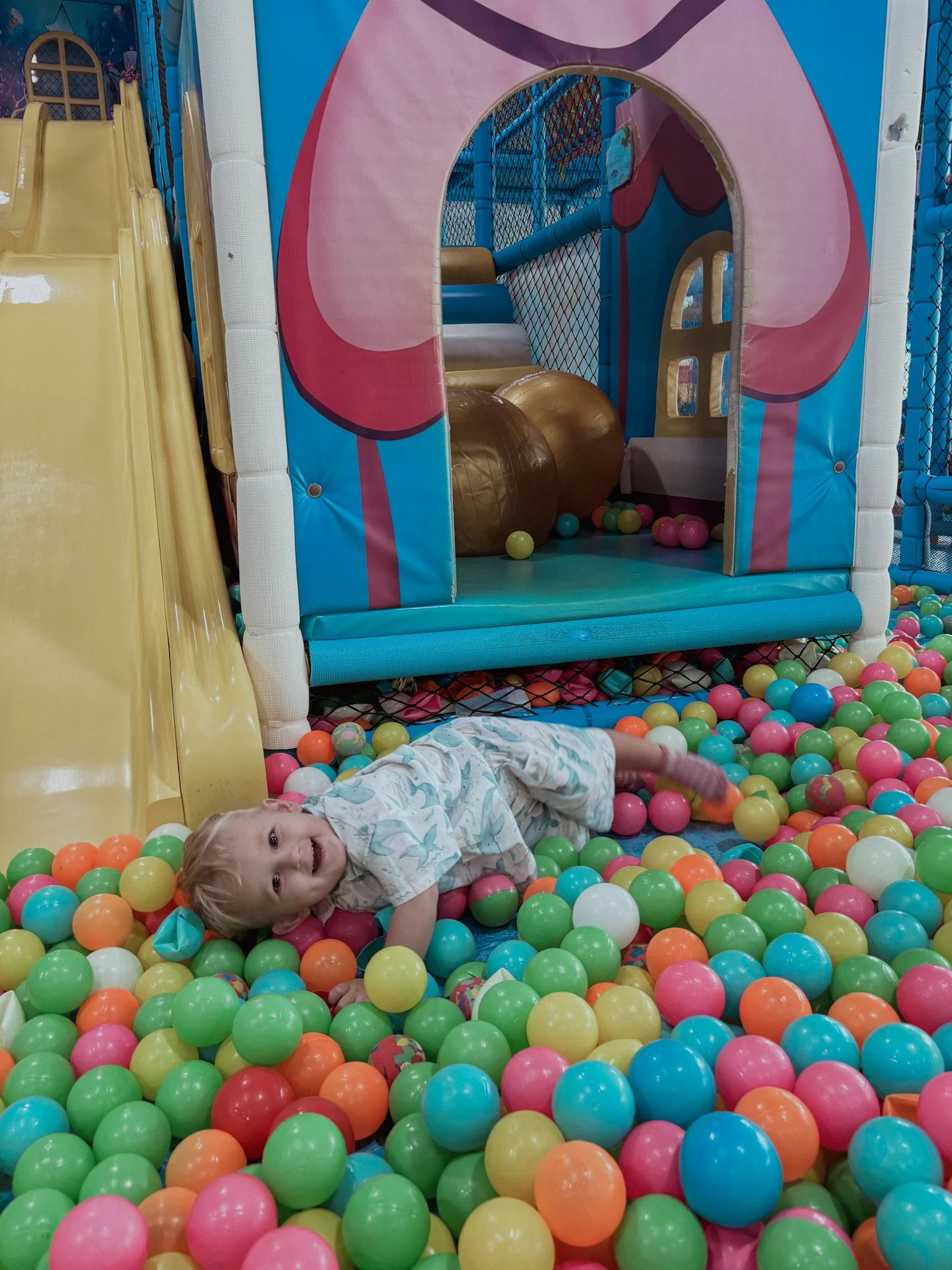 A young child lying on a colorful ball pit surrounded by plastic balls of various colors, inside an indoor play area with a slide and soft play structures.