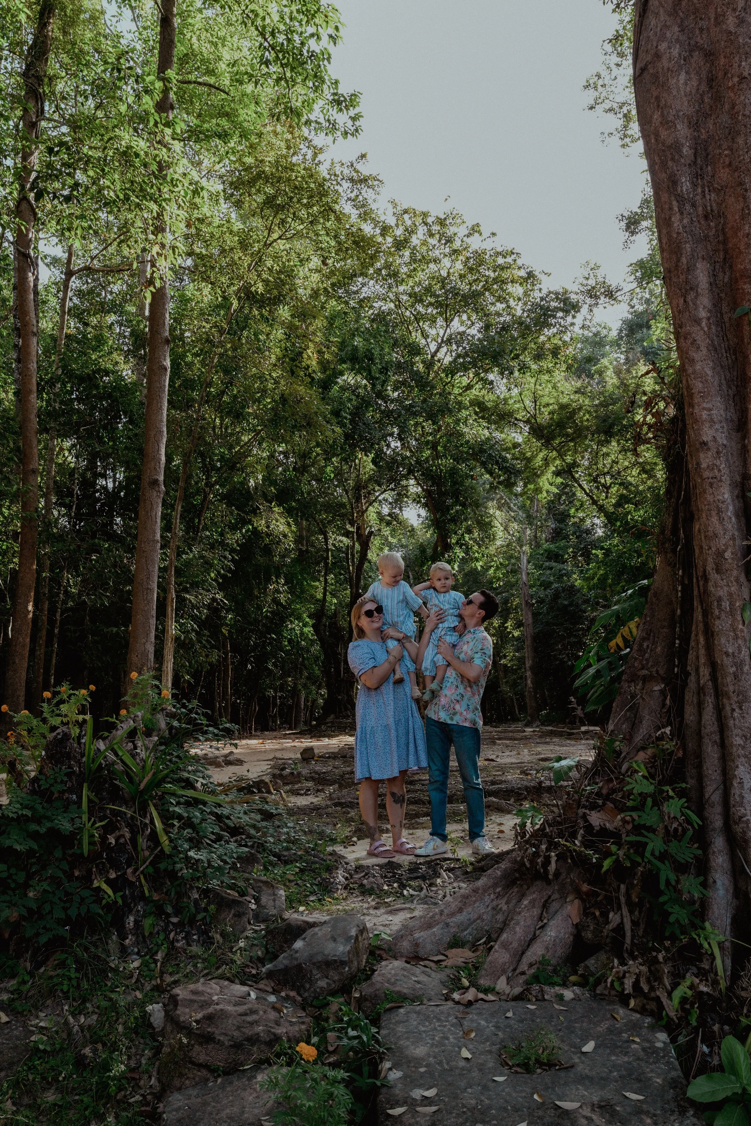 A woman and man are standing in a forest, each holding a child. The children are dressed in matching blue outfits. The woman is wearing sunglasses and a blue dress, and the man is wearing a patterned shirt and jeans. They are smiling and looking at each other amid greenery and tall trees.