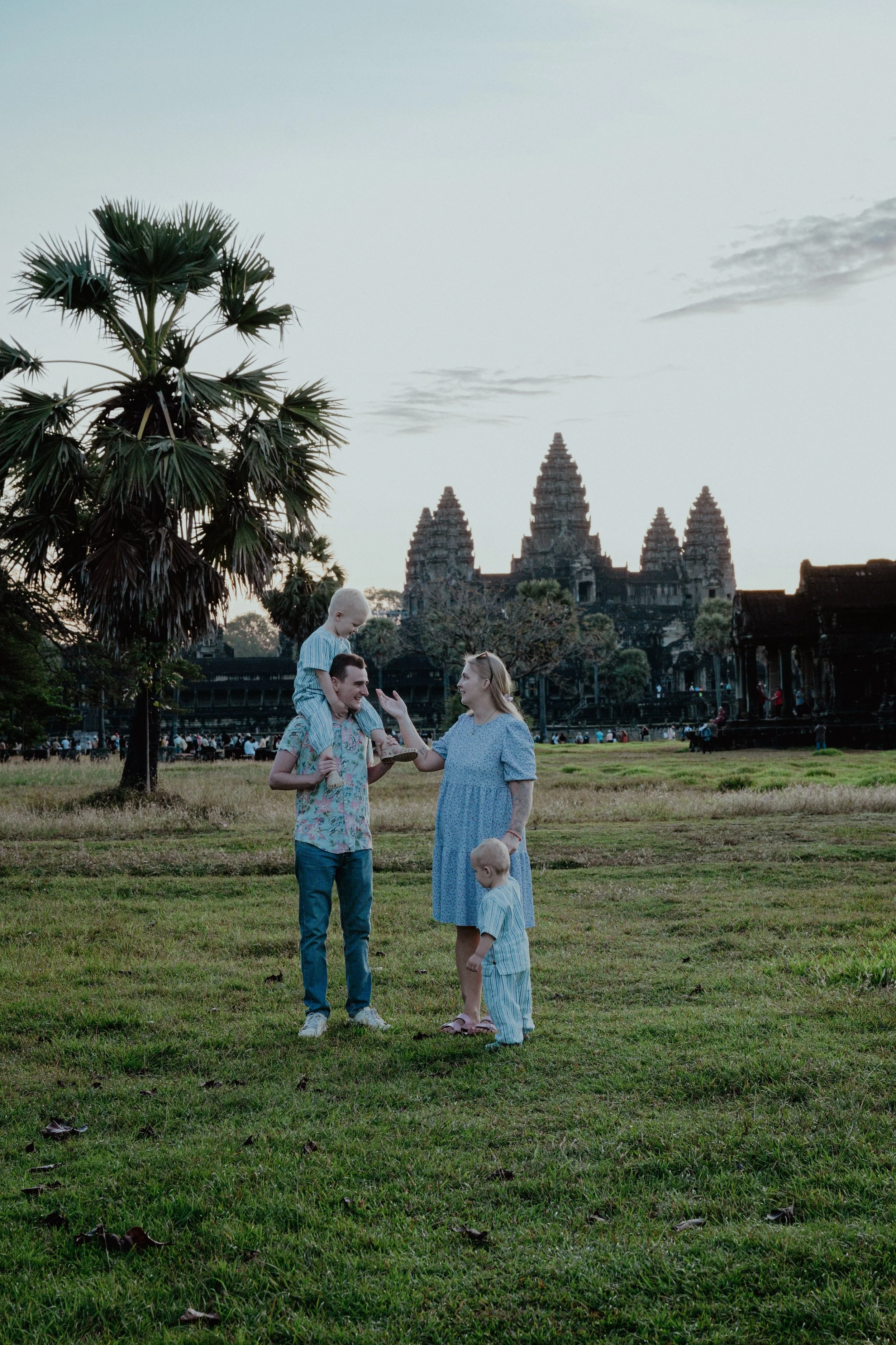 A family of four enjoying a moment on a grassy field near Angkor Wat at sunset. The father carries a child on his shoulders, the mother stands with her hand raised, and a young child stands nearby. Lush trees and Angkor Wat's temples are visible in the background.