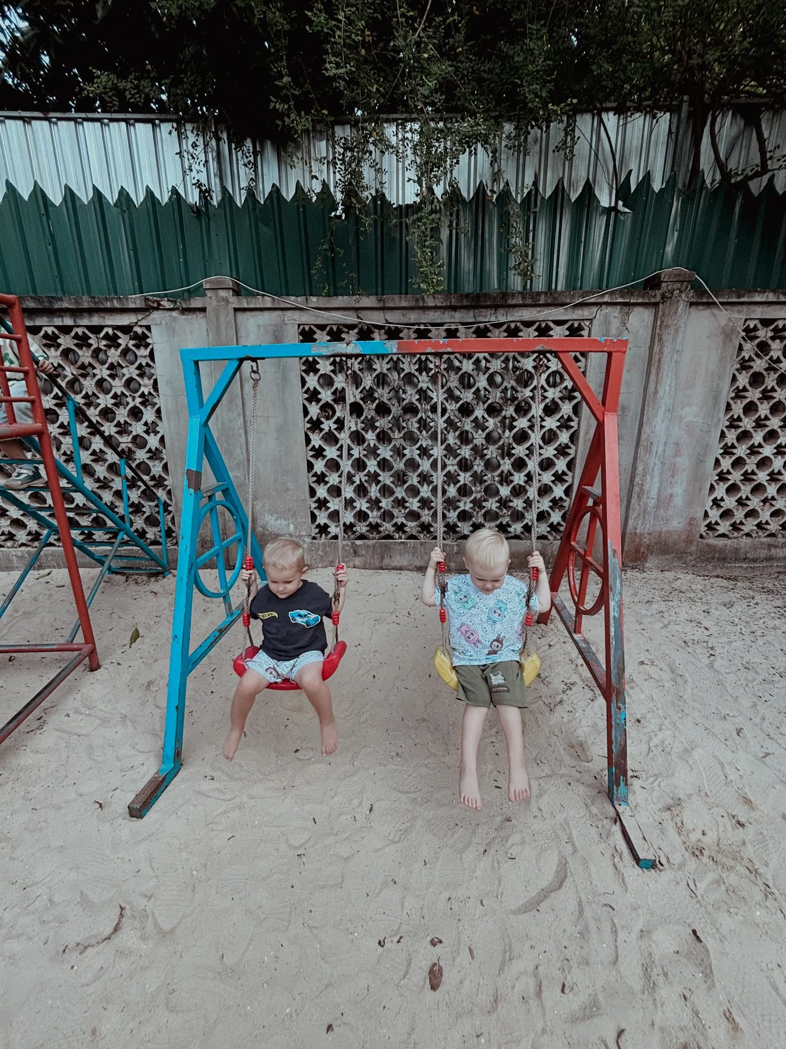 Two young boys sitting on swings at a playground with a sandy ground, surrounded by a concrete fence and green plants.