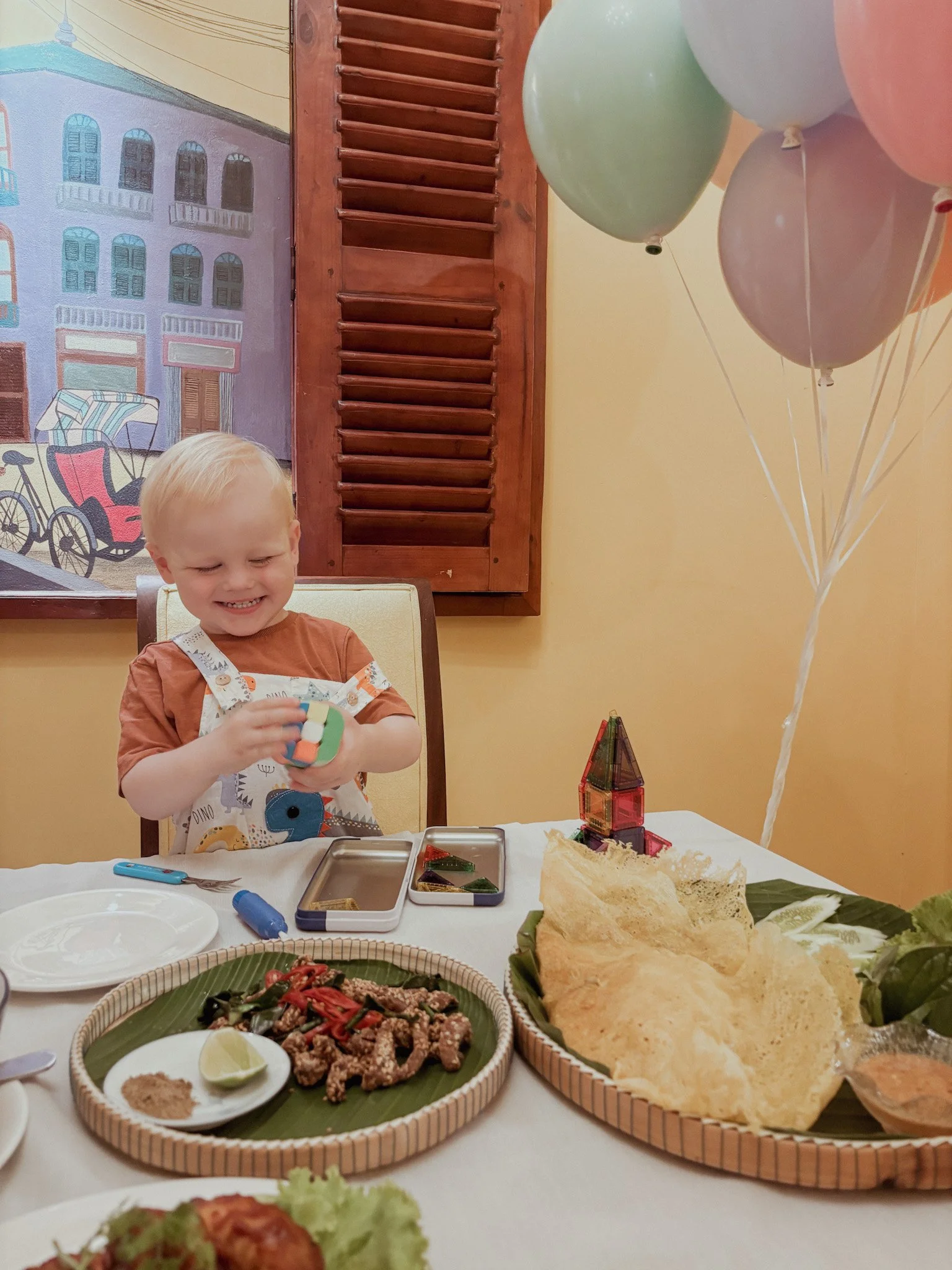 A young boy with blonde hair, wearing a brown shirt and a colorful bib, smiling and playing with a toy at a birthday celebration. There are balloons, a tray of food including crispy chips and grilled meat, and a decorative background with a painting and wooden shutter.