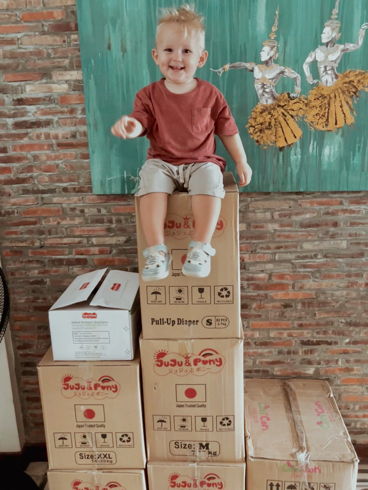 A young boy sitting on top of stacked cardboard boxes, smiling and pointing. The background features a brick wall and a painted mural of dancers.