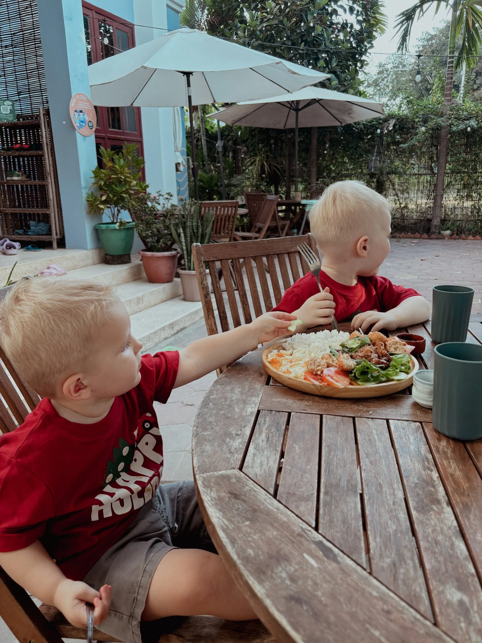Two young boys with blond hair wearing red shirts, enjoying a meal outdoors at a wooden table with a pizza on a tray, surrounded by cups and plates, in a backyard patio with green plants, potted plants, umbrellas, and other outdoor furniture.