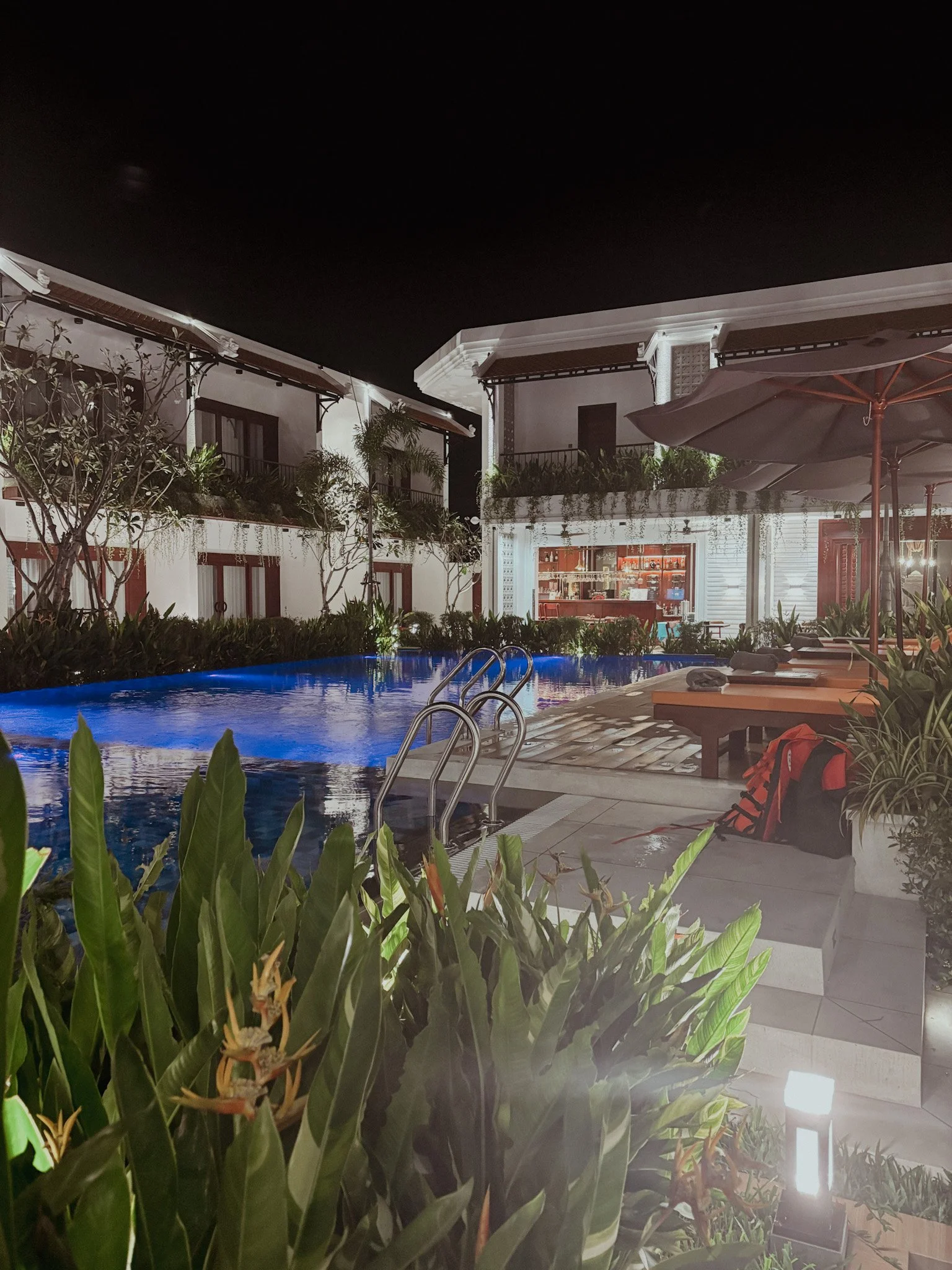 Nighttime view of a hotel pool area with lounge chairs, potted plants, and a bar area in the background.