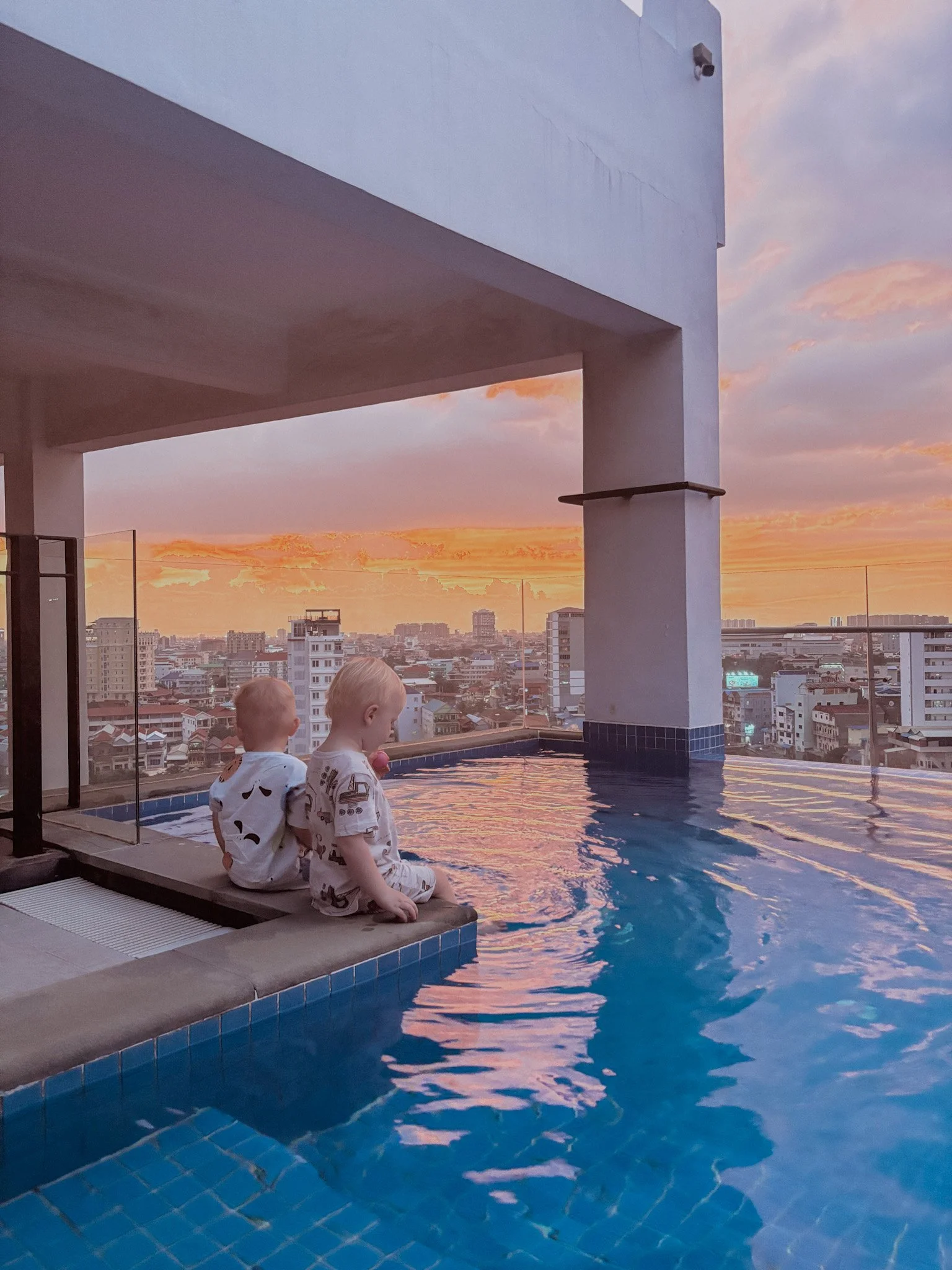 Two young children sitting on the edge of an infinity pool on a rooftop, overlooking a cityscape at sunset.
