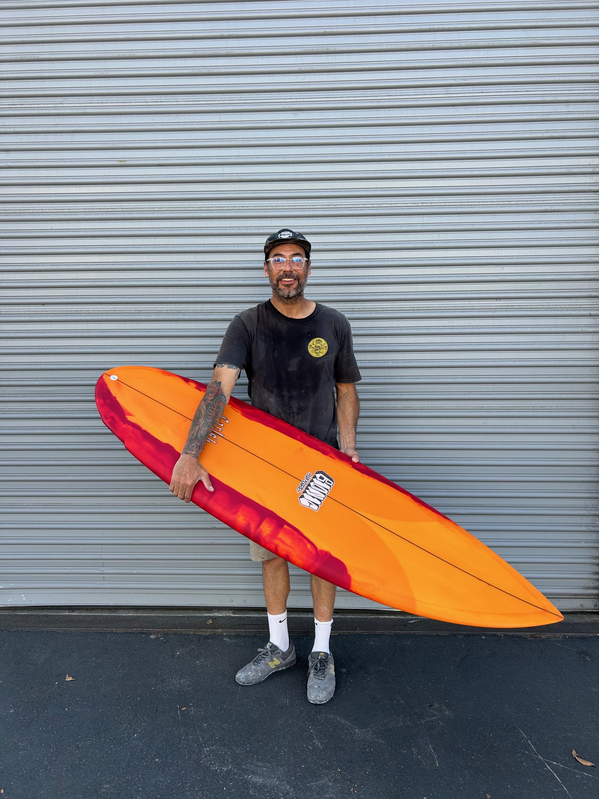 A man standing in front of a metal garage door, holding an orange and red surfboard. He is wearing a black T-shirt, shorts, gray sneakers, and a black cap. He has glasses and a tattooed arm, smiling at the camera.