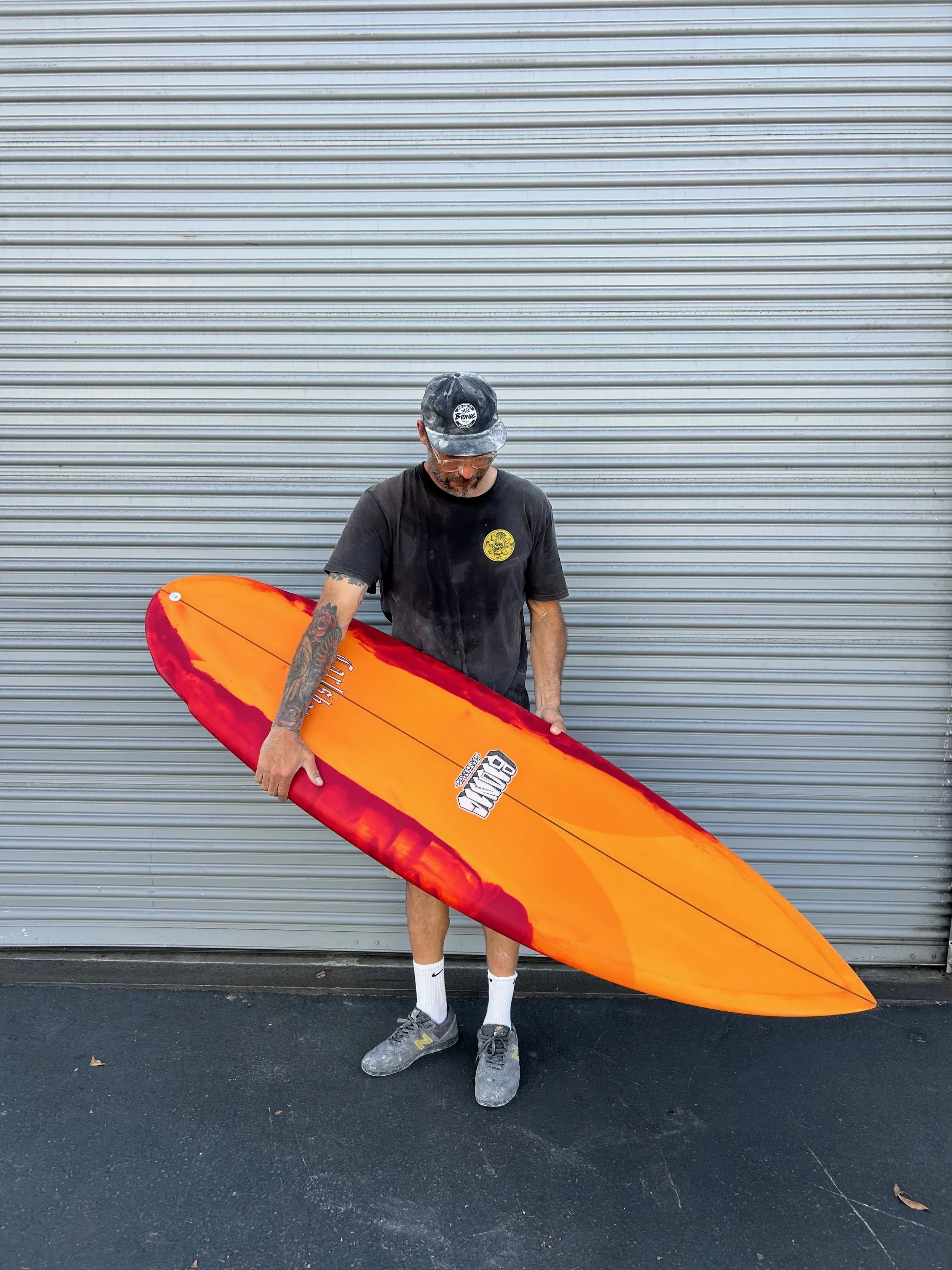 A man wearing a black t-shirt, gray sneakers, and white socks, holding an orange and red surfboard, standing in front of a gray metal roll-up door.