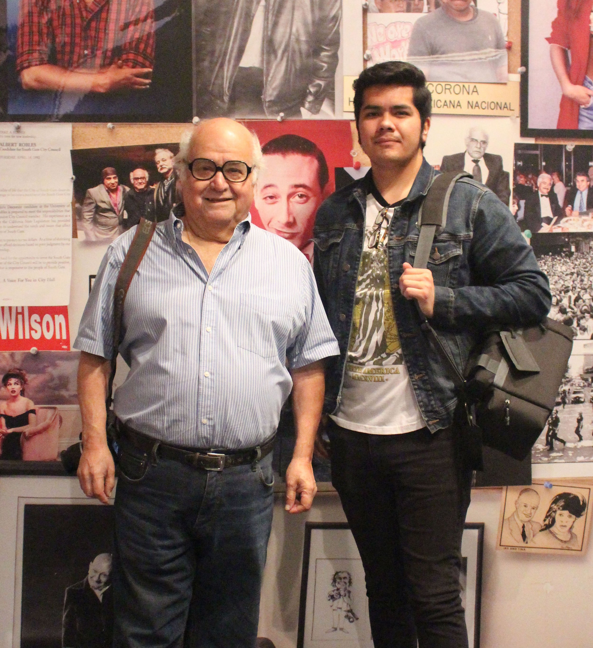 Two men standing in front of a collage wall of photographs and posters, one older with glasses and a striped shirt, the other younger with a denim jacket and backpack.