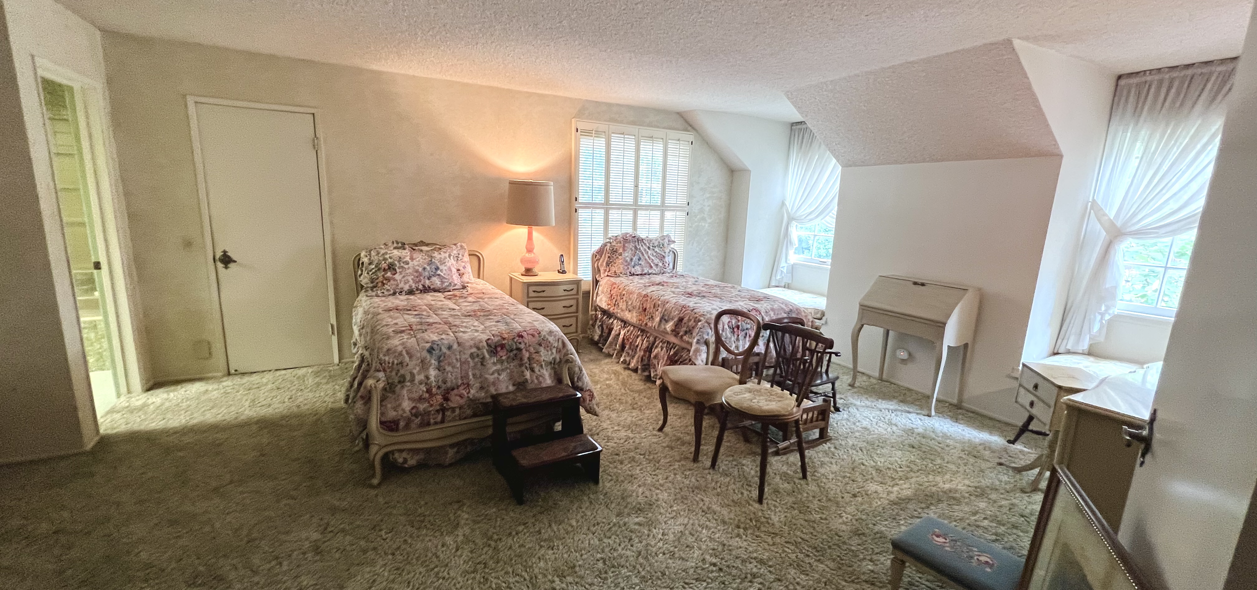 A bedroom with two twin beds with floral bedding, a nightstand with a lamp, and multiple chairs of different styles, with large windows with white curtains and beige carpeting.