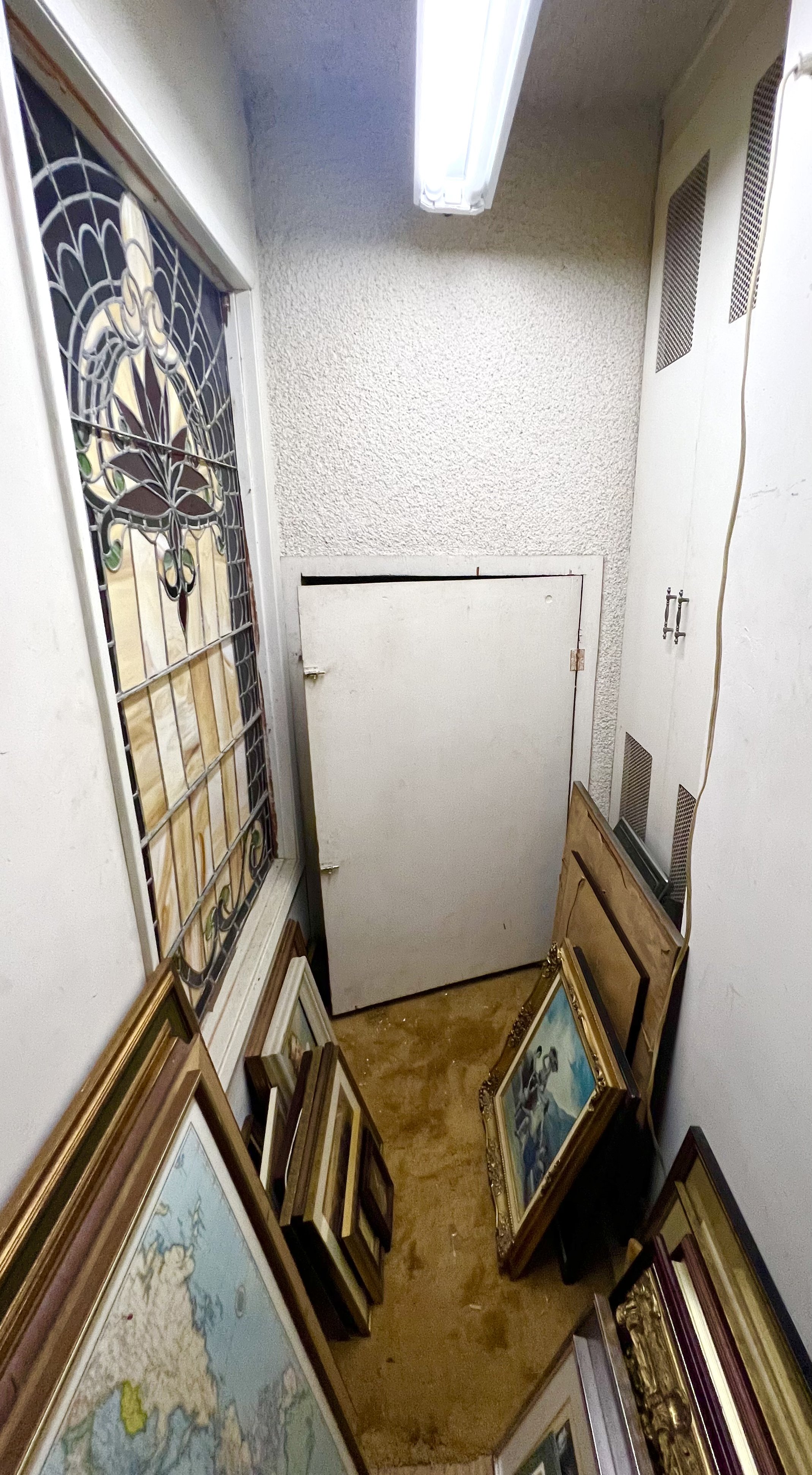 Small room with framed pictures and artwork leaning against the walls, a stained glass window, and a door leading to an attic or crawl space.