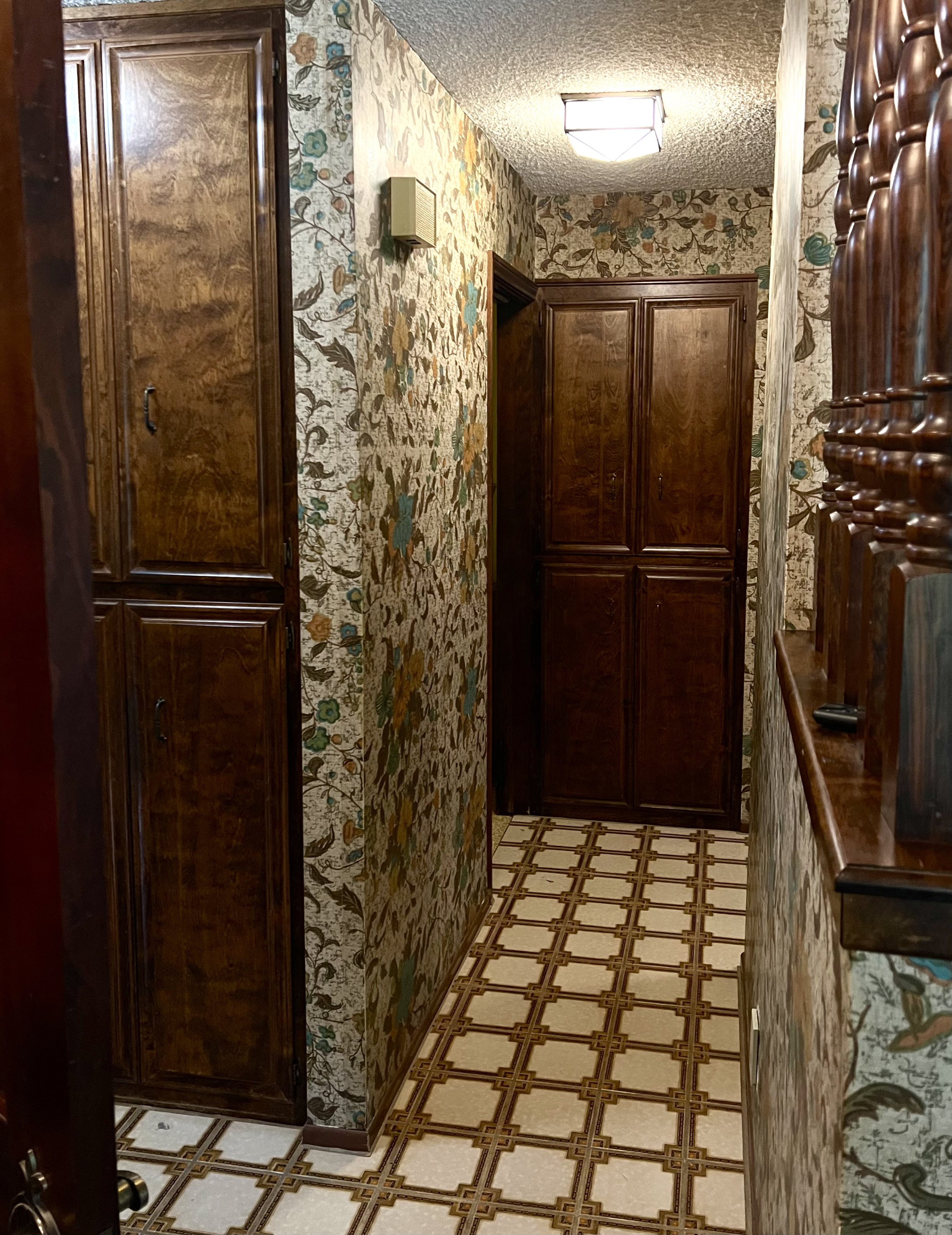 Narrow hallway with floral wallpaper, wooden cabinets, and patterned tile flooring, illuminated by ceiling light.
