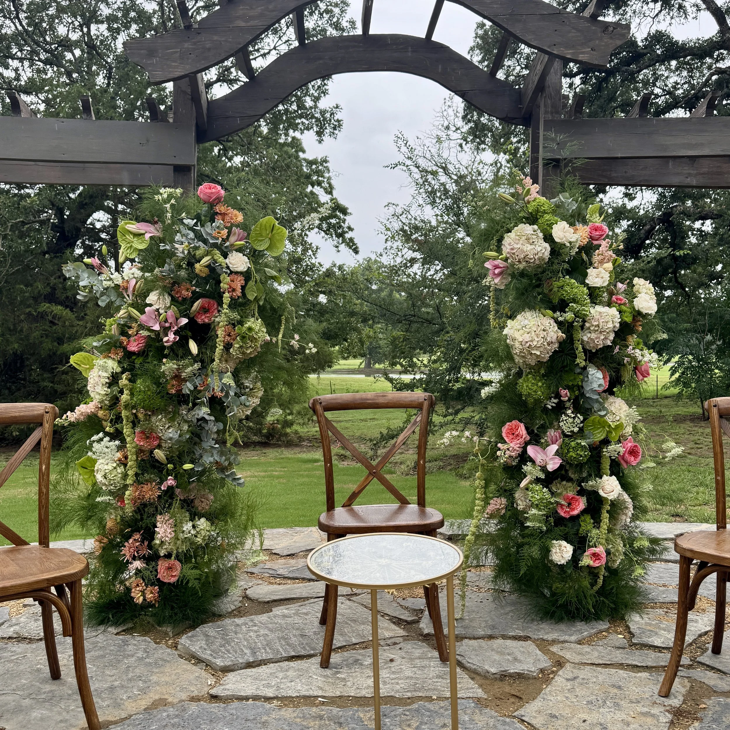 Wedding ceremony setup with floral arrangements and chairs under a wooden arch outside on a stone patio.