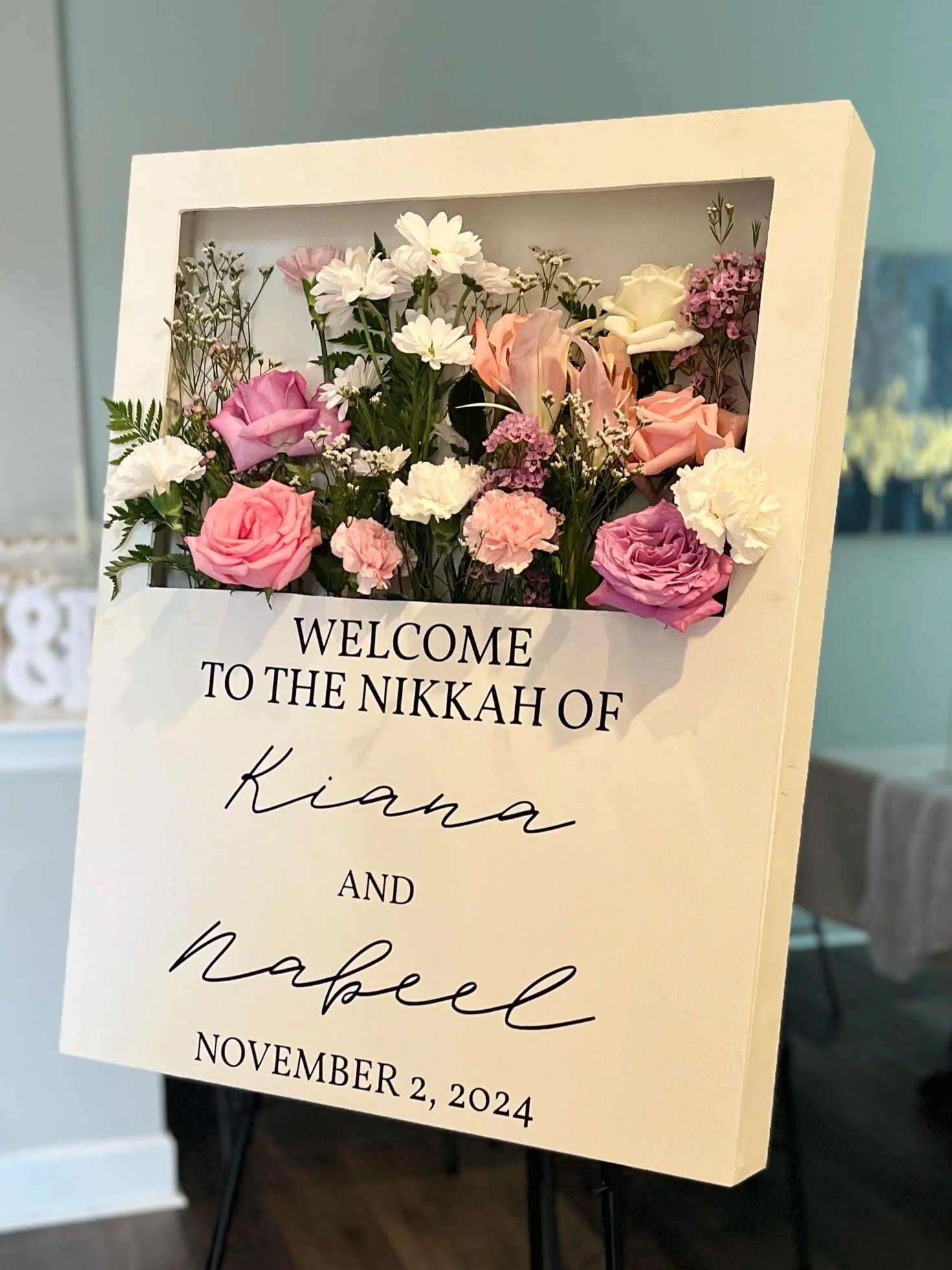 A floral welcome sign with pink, white, and purple flowers in a display case, welcoming Kiana and Nabeel to their nikkah on November 2, 2024.