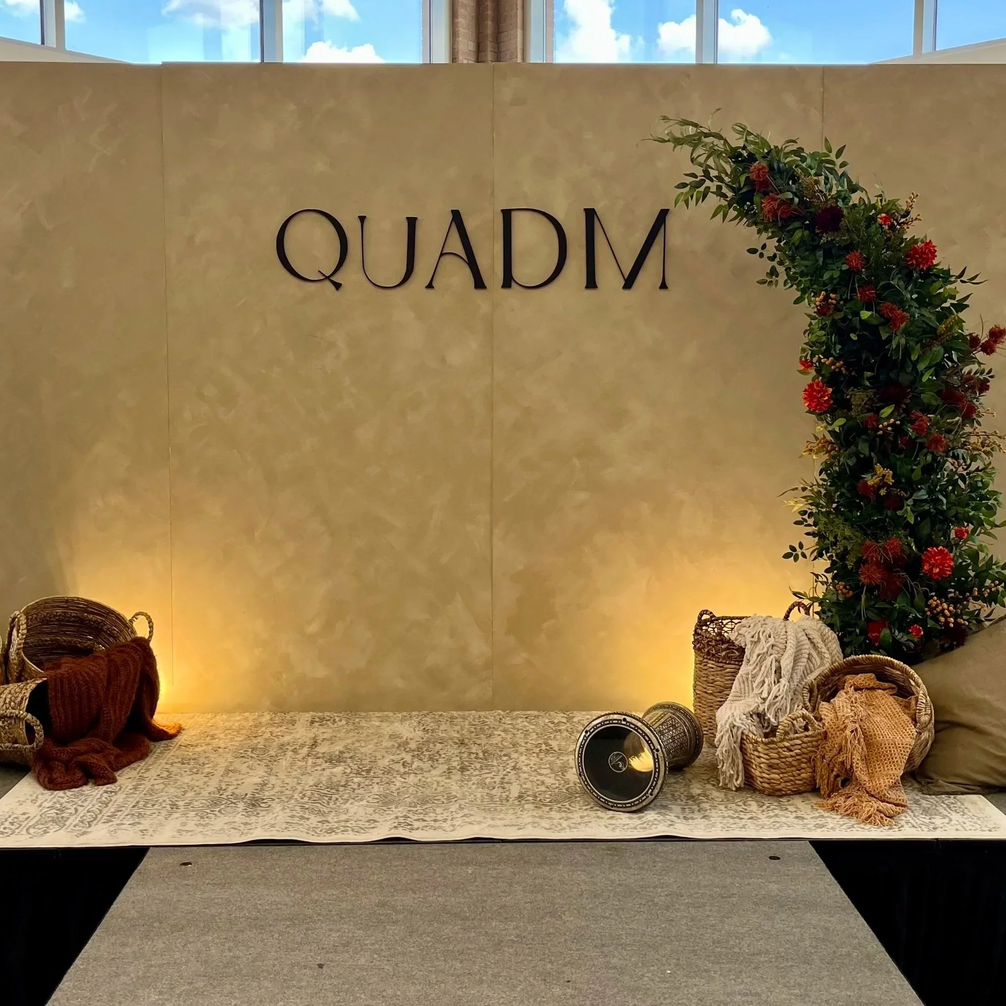 A decorated display table with baskets, blankets, and a pillow in front of a beige wall with 'QUADM' lettering and a floral arrangement on the right side.