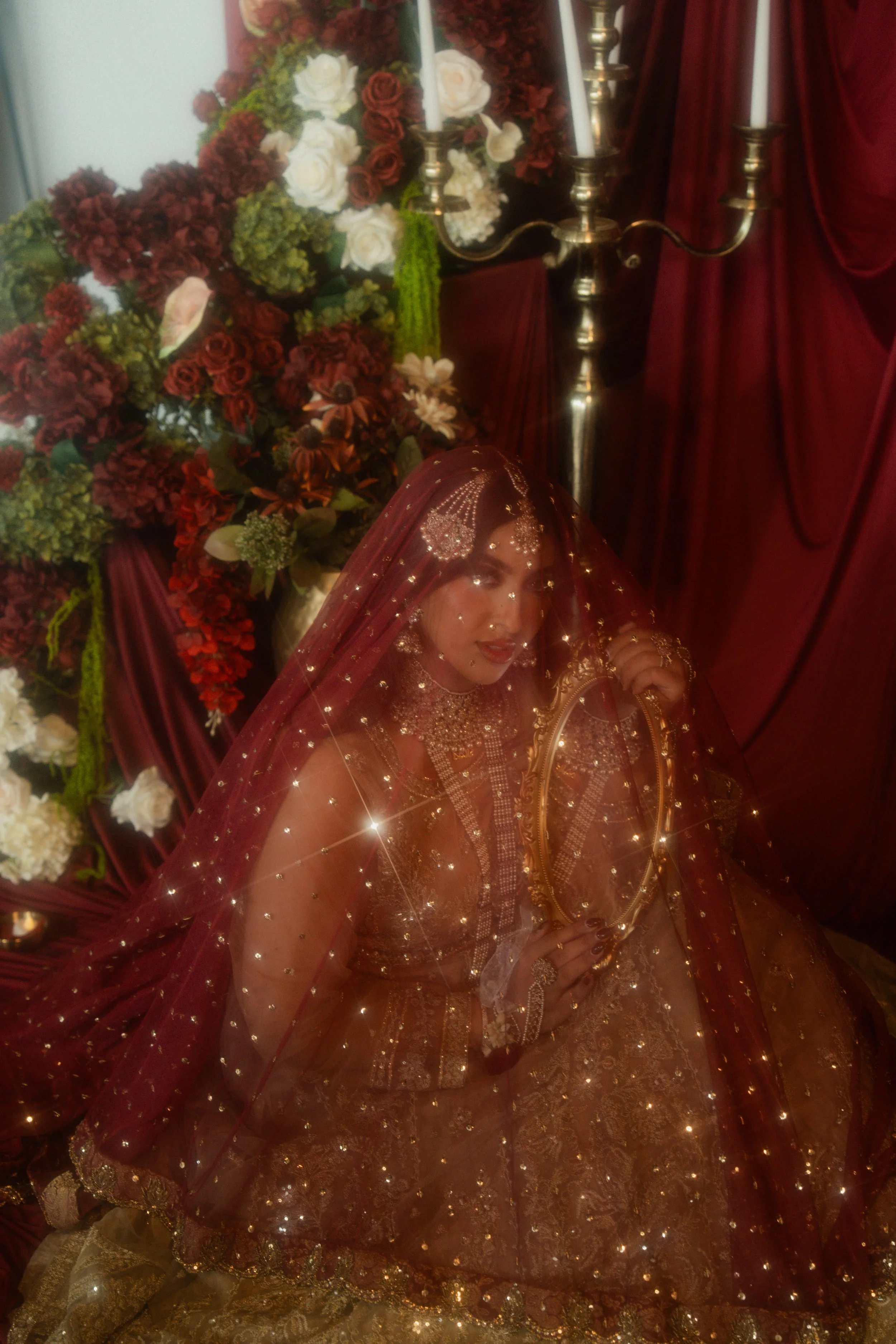 A woman dressed in an elaborate, glittering maroon and gold outfit, wearing jewelry, holding a mirror, and sitting in front of a large floral arrangement with red, white, and green flowers, with a red curtain in the background.