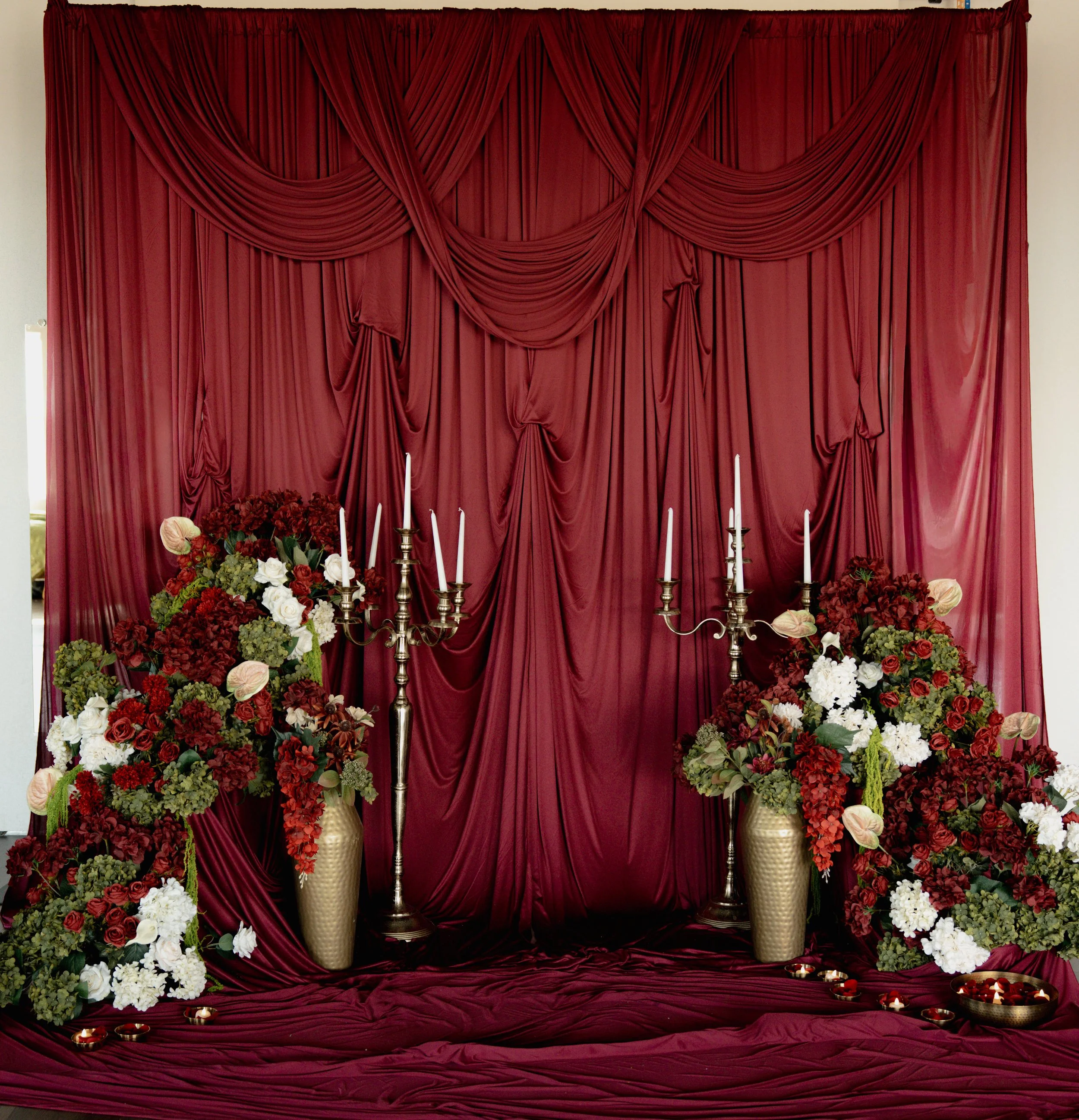 Red draped fabric backdrop with large floral arrangements in gold vases, silver candelabras with white candles, and scattered candles on the floor, creating an elegant setting.