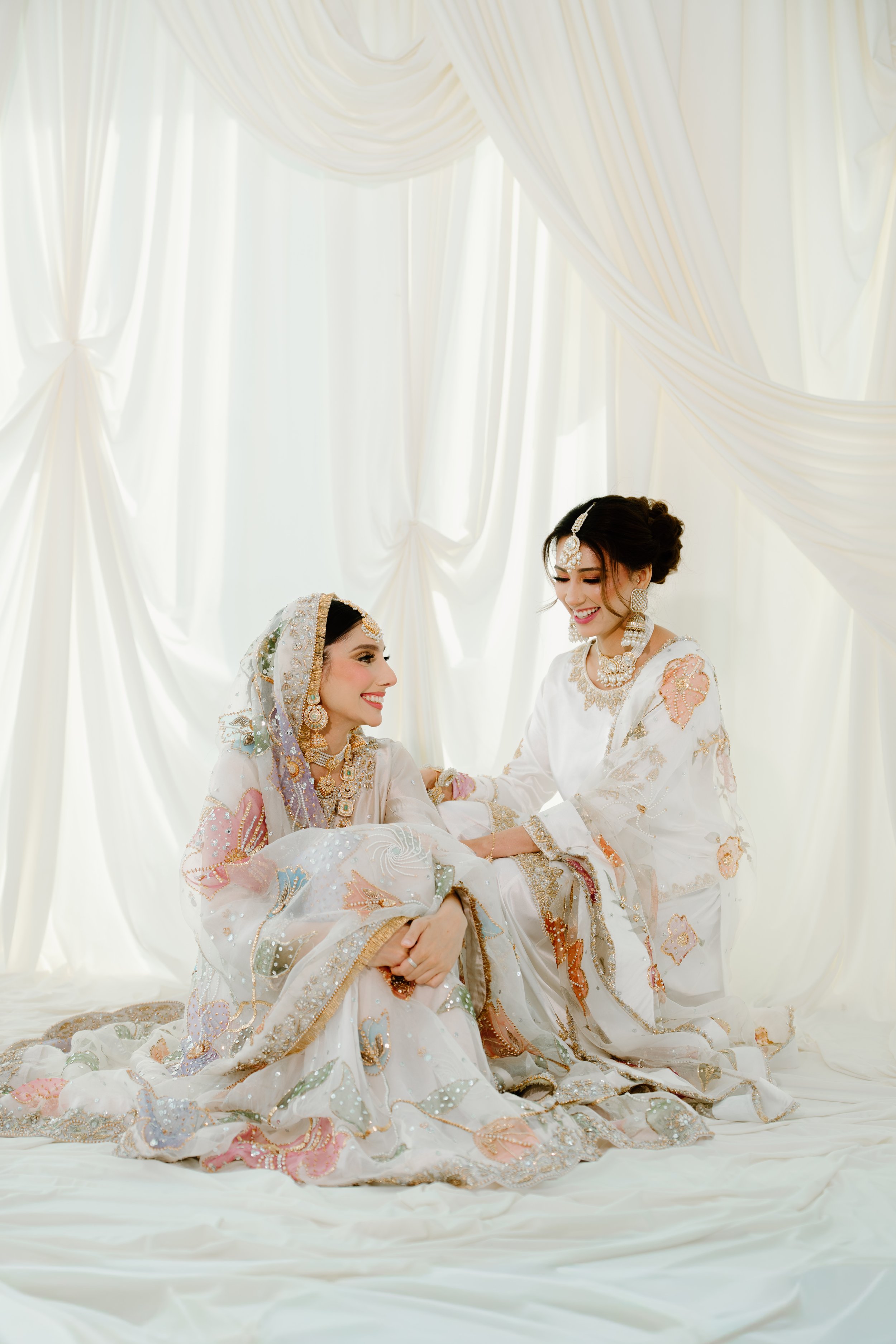 Two women dressed in traditional South Asian wedding attire, nikkah dress, sitting on a white fabric floor in front of a white draped curtain, smiling and holding hands during a wedding celebration.
