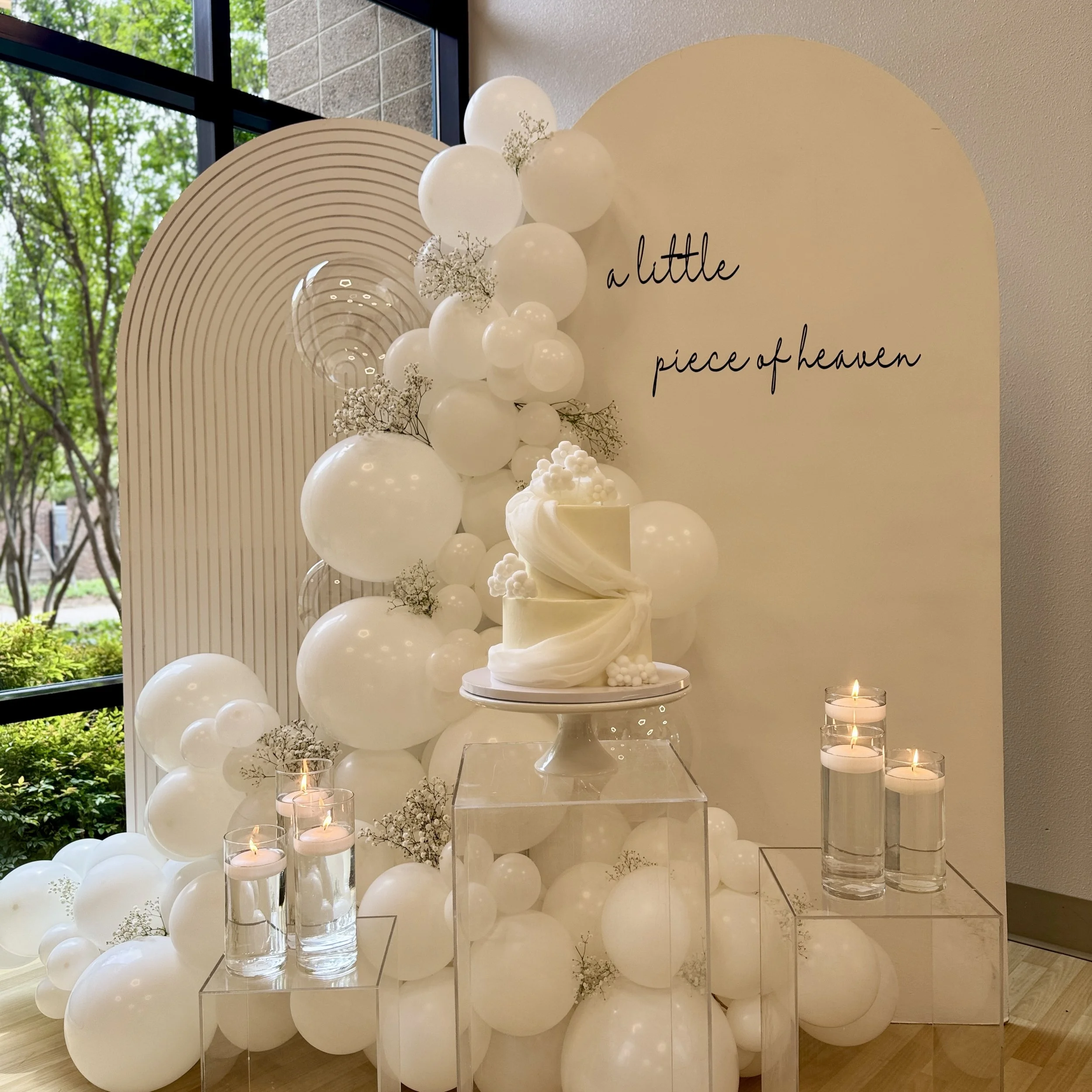Elegant celebration display with white balloons, a white cake, candles, and a sign that reads 'a little piece of heaven'.