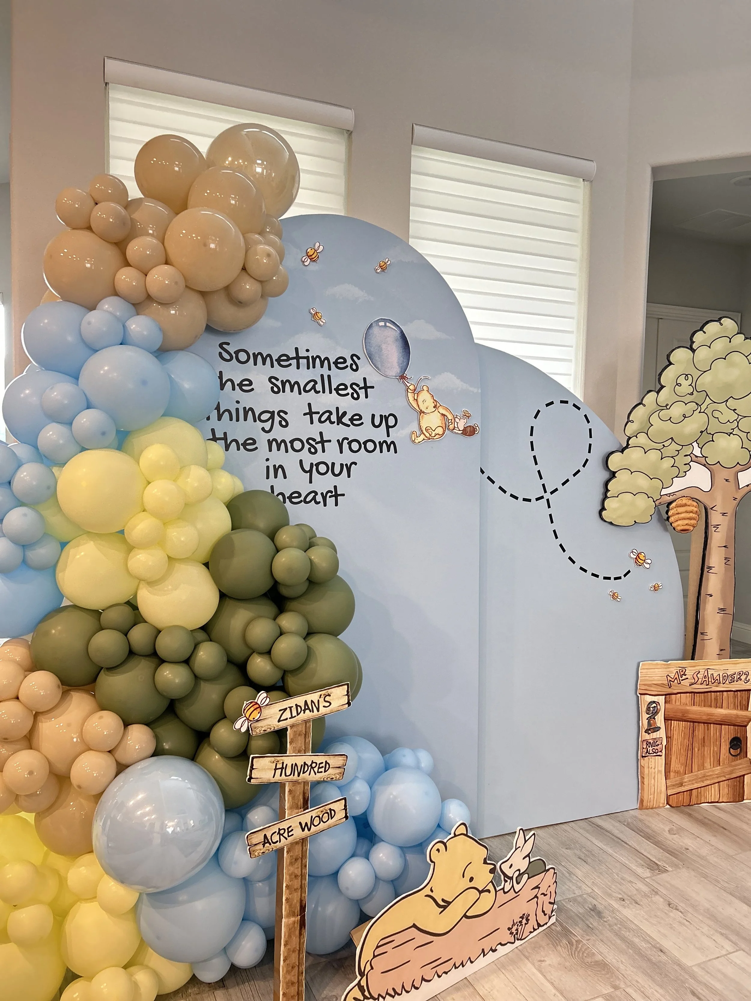 Decorative setup with a Winnie the Pooh theme, featuring a large tree with balloons, a signpost reading ‘Zidan’s Hundred Acre Wood’, a cutout of Winnie the Pooh and Piglet, and a backdrop with a quote about small things taking up room in your heart.