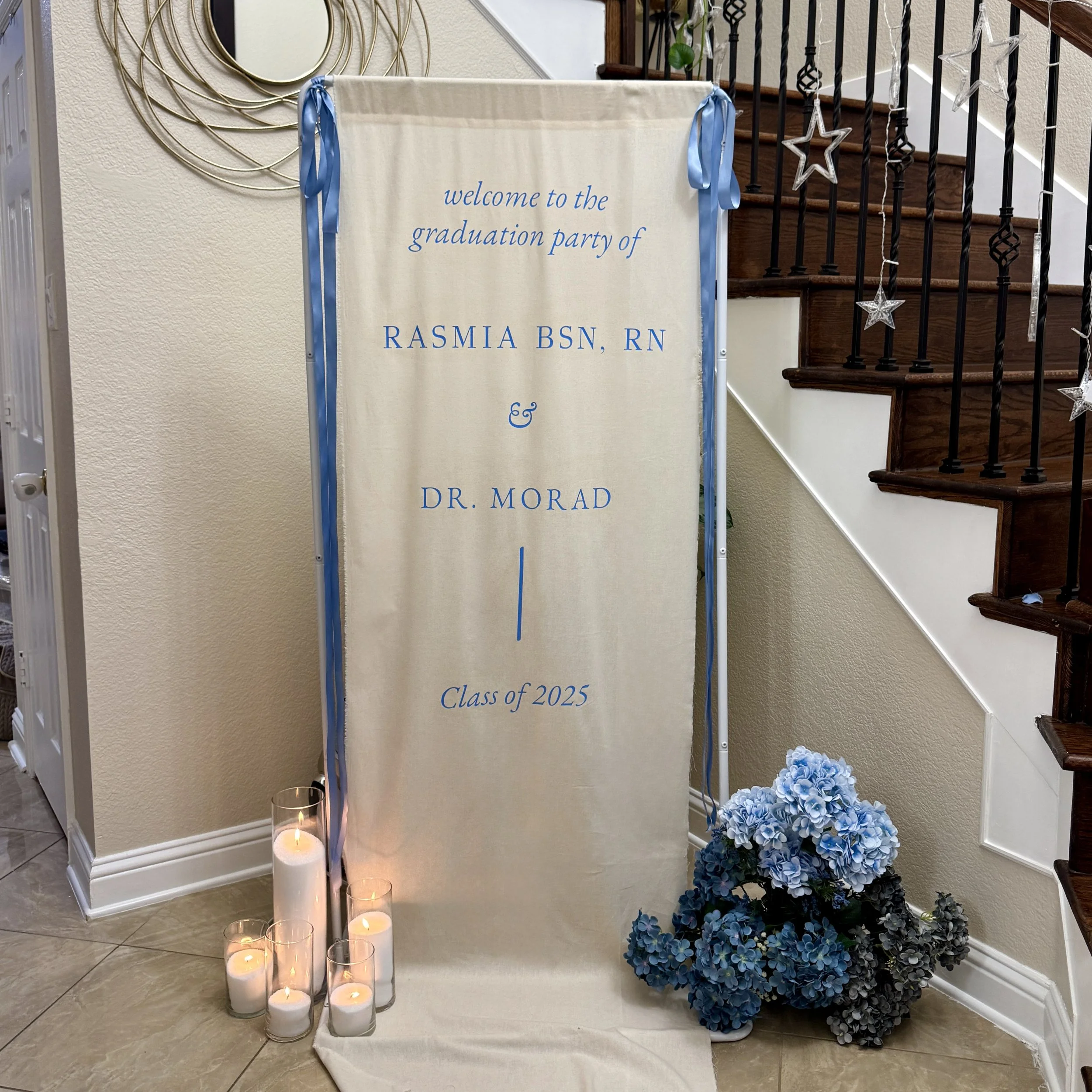 A beige fabric backdrop for a graduation party with blue ribbons on each side. The backdrop displays text welcoming guests to the graduation party of Rasmia BSN, RN, and Dr. Morad, for the class of 2025. There are lit white candles in glass holders o
