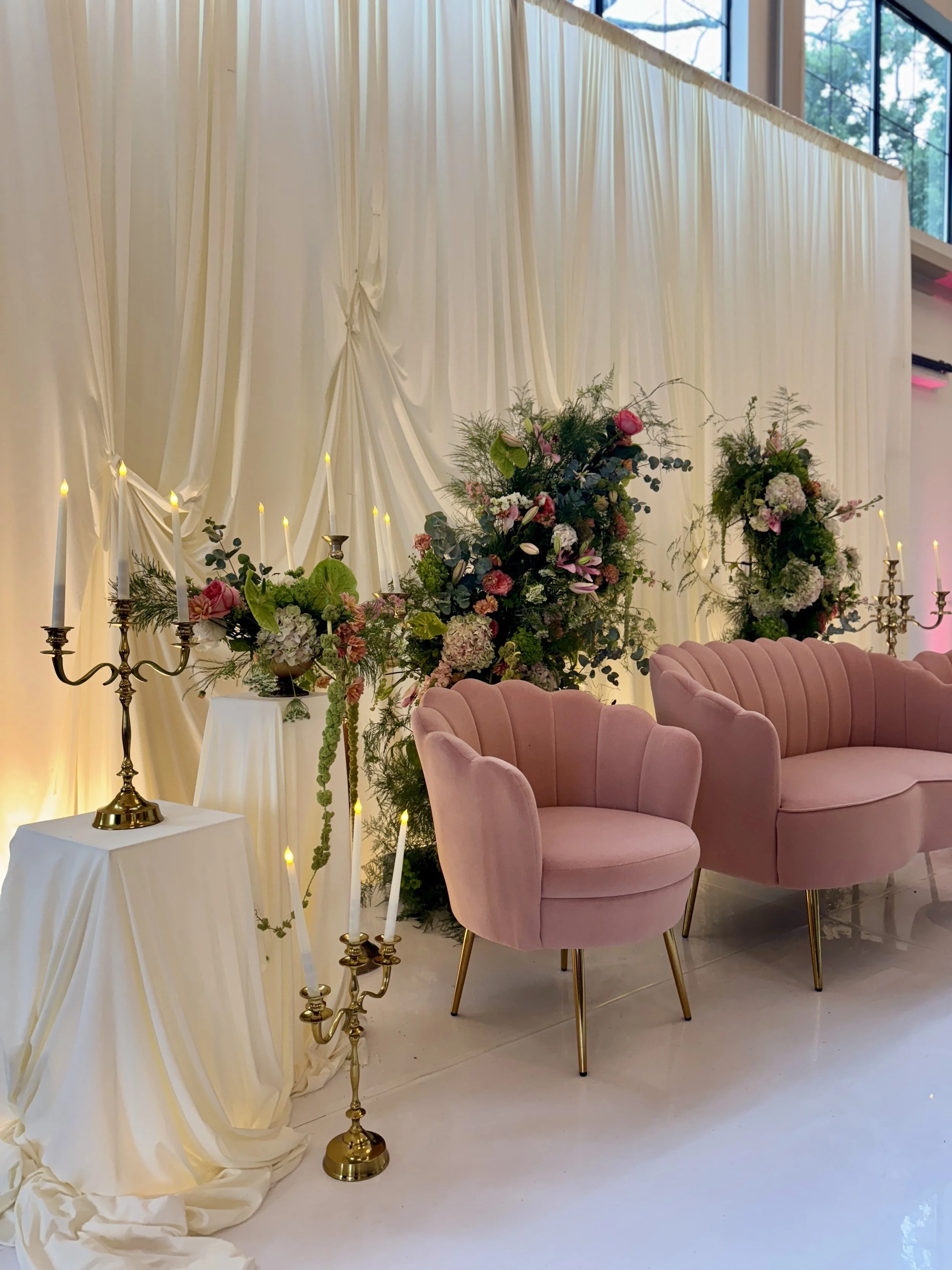 Elegant wedding or event setup with pink velvet armchairs, floral arrangements, white drapery backdrop, and gold candelabras with lit candles.