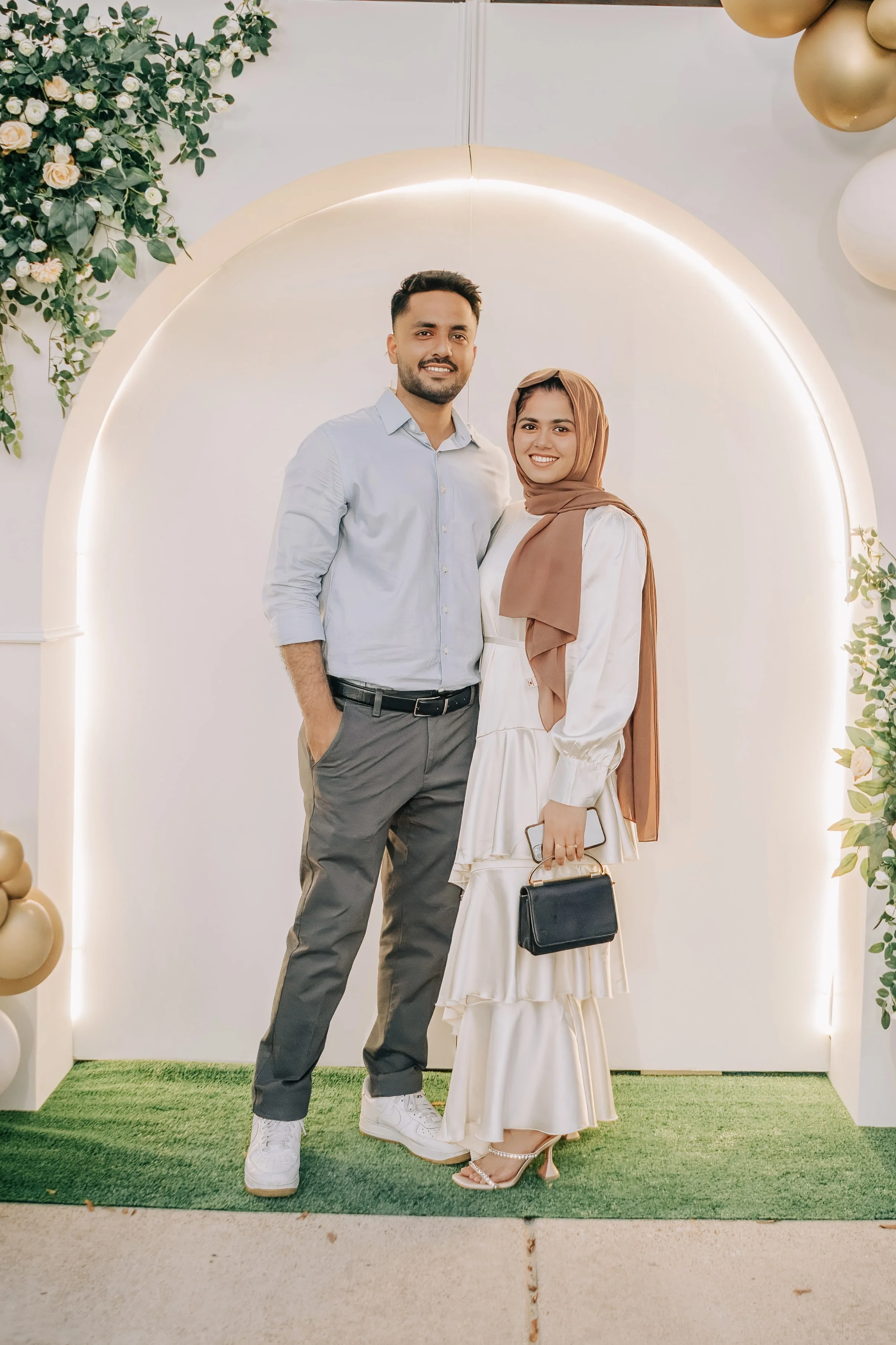 A man and woman smiling and standing together in front of a white backdrop with decorative balloons and greenery. The man is wearing a light blue shirt, gray pants, and white sneakers. The woman is wearing a white dress, a brown headscarf, and heels, and holding a black handbag.