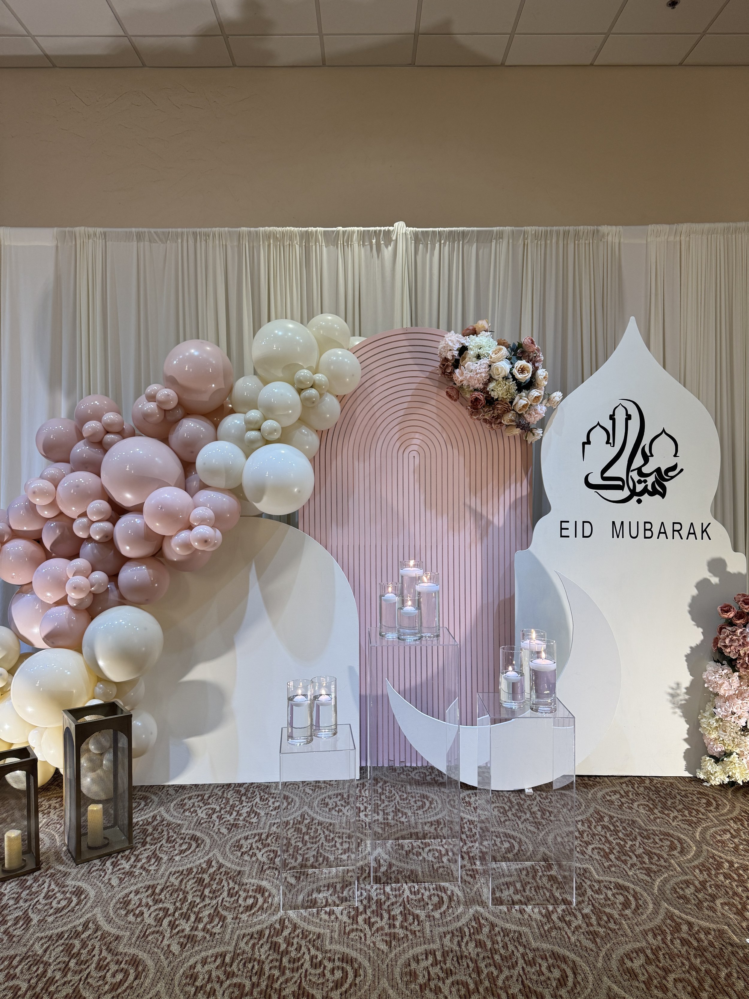 Eid Mubarak greeting with decorative backdrop featuring pink and white balloons, floral arrangements, candles in glass holders, and a white panel with Arabic calligraphy.