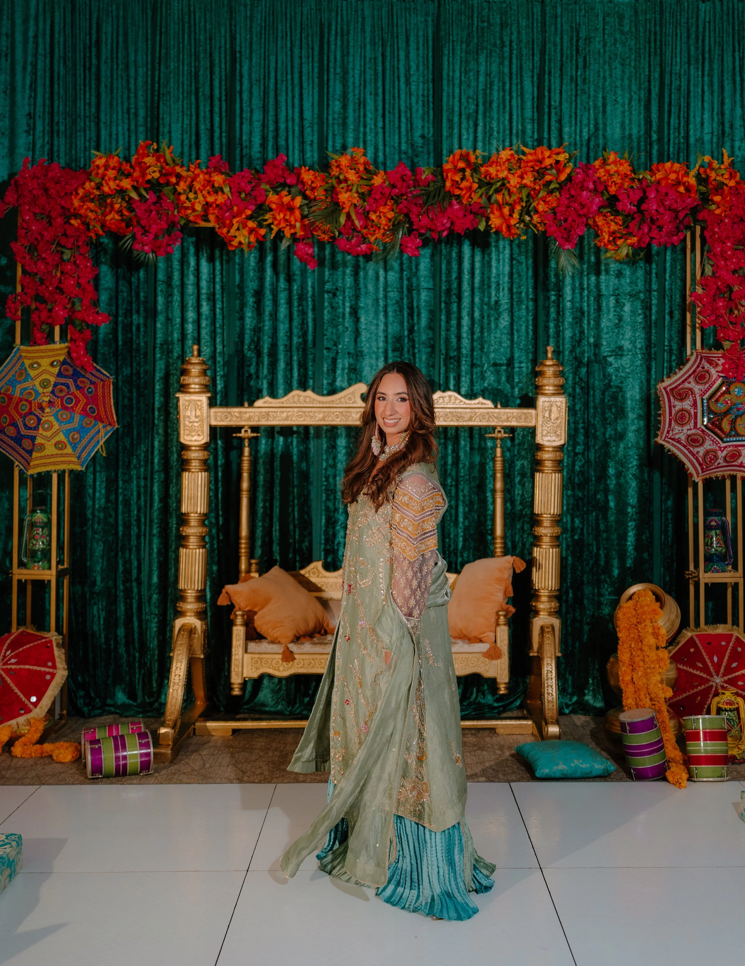 A woman in traditional attire standing in front of a decorated backdrop with colorful umbrellas, drums, and floral arrangements at a cultural celebration or event.