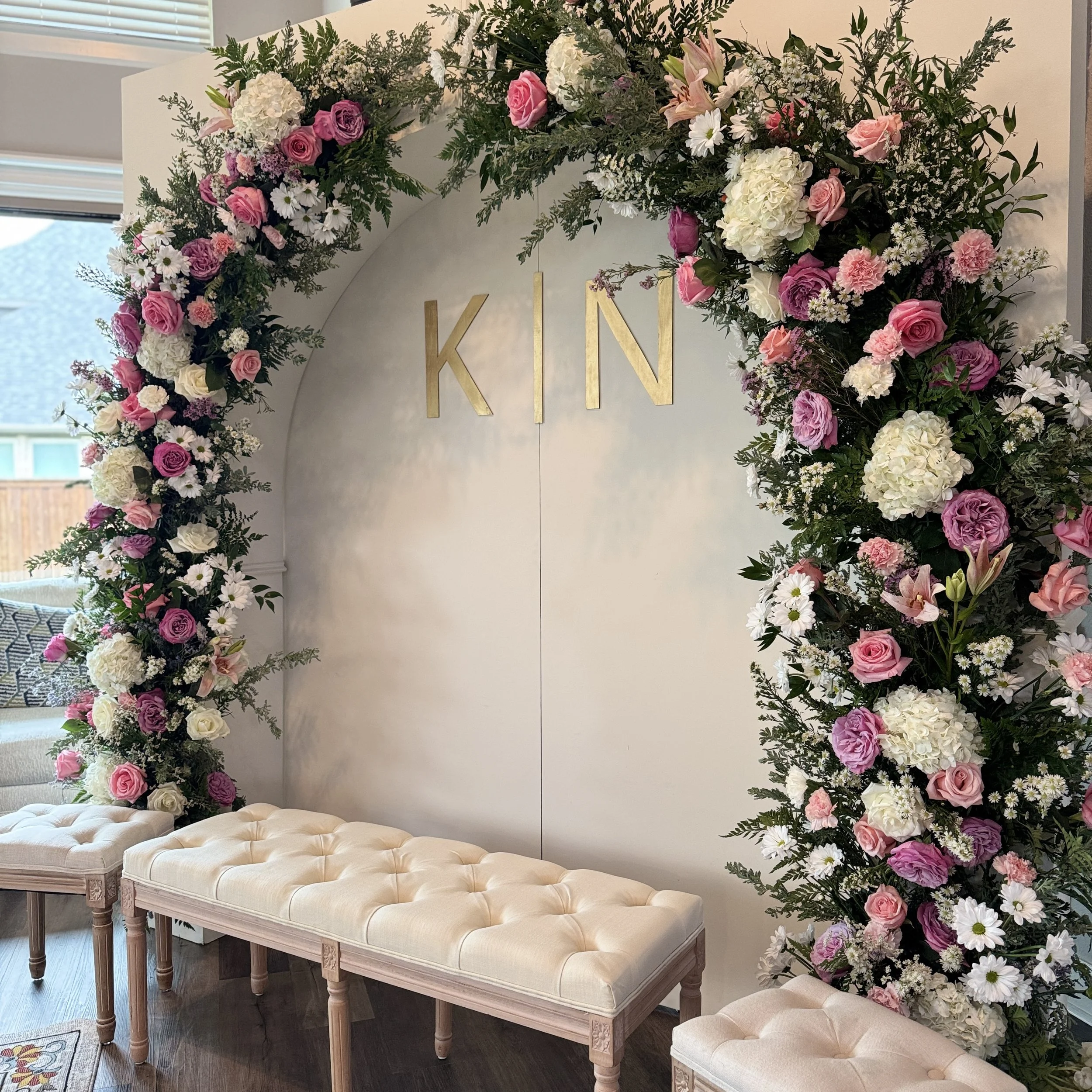 A floral arch with pink, white, and purple flowers surrounding a white backdrop with the gold letters 'K' and 'N' hanging in the center, and beige tufted benches in front.