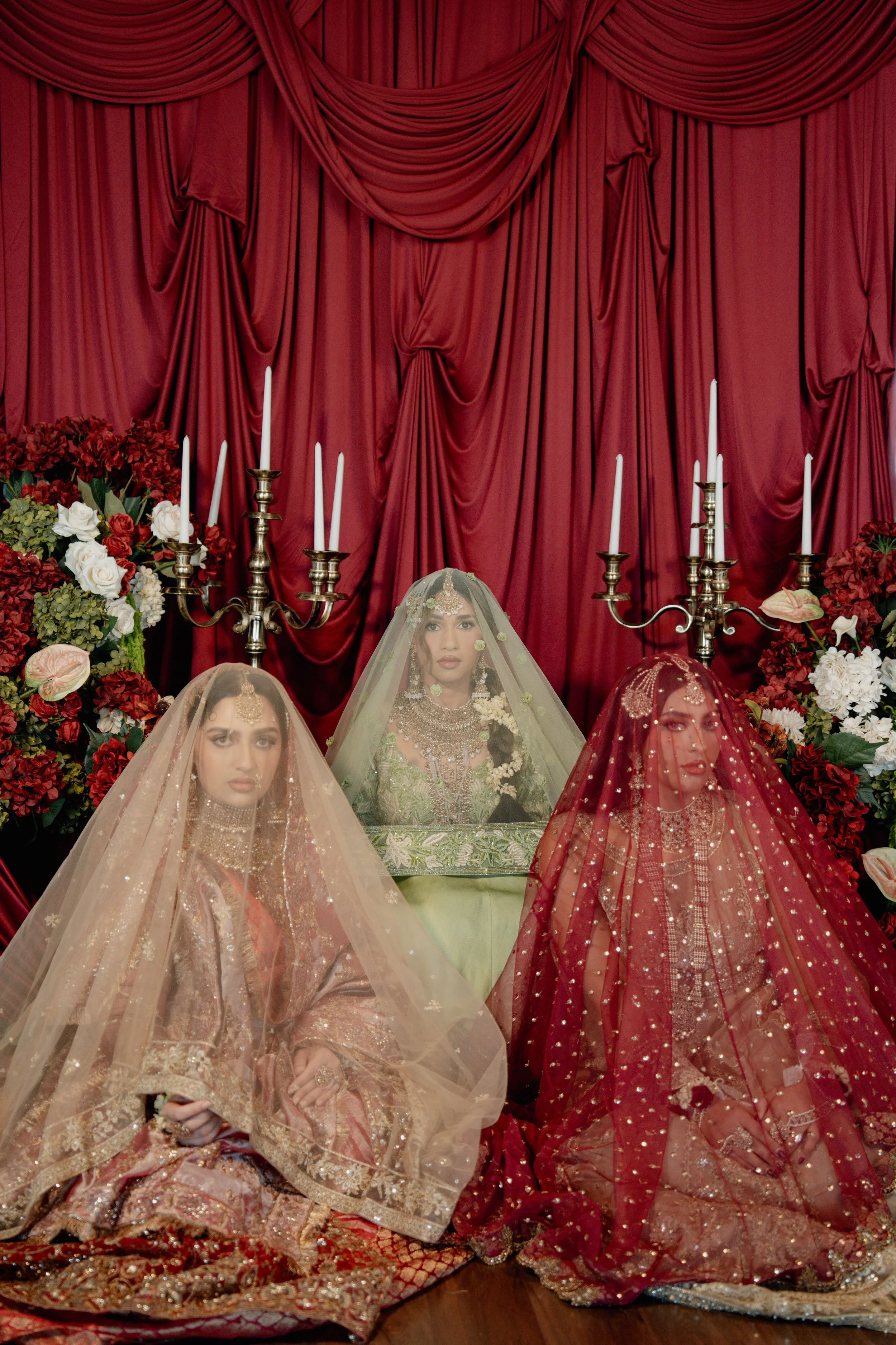 Three women dressed in traditional South Asian bridal attire, seated in front of a red curtain with floral arrangements and candelabras. Luxury desi wedding