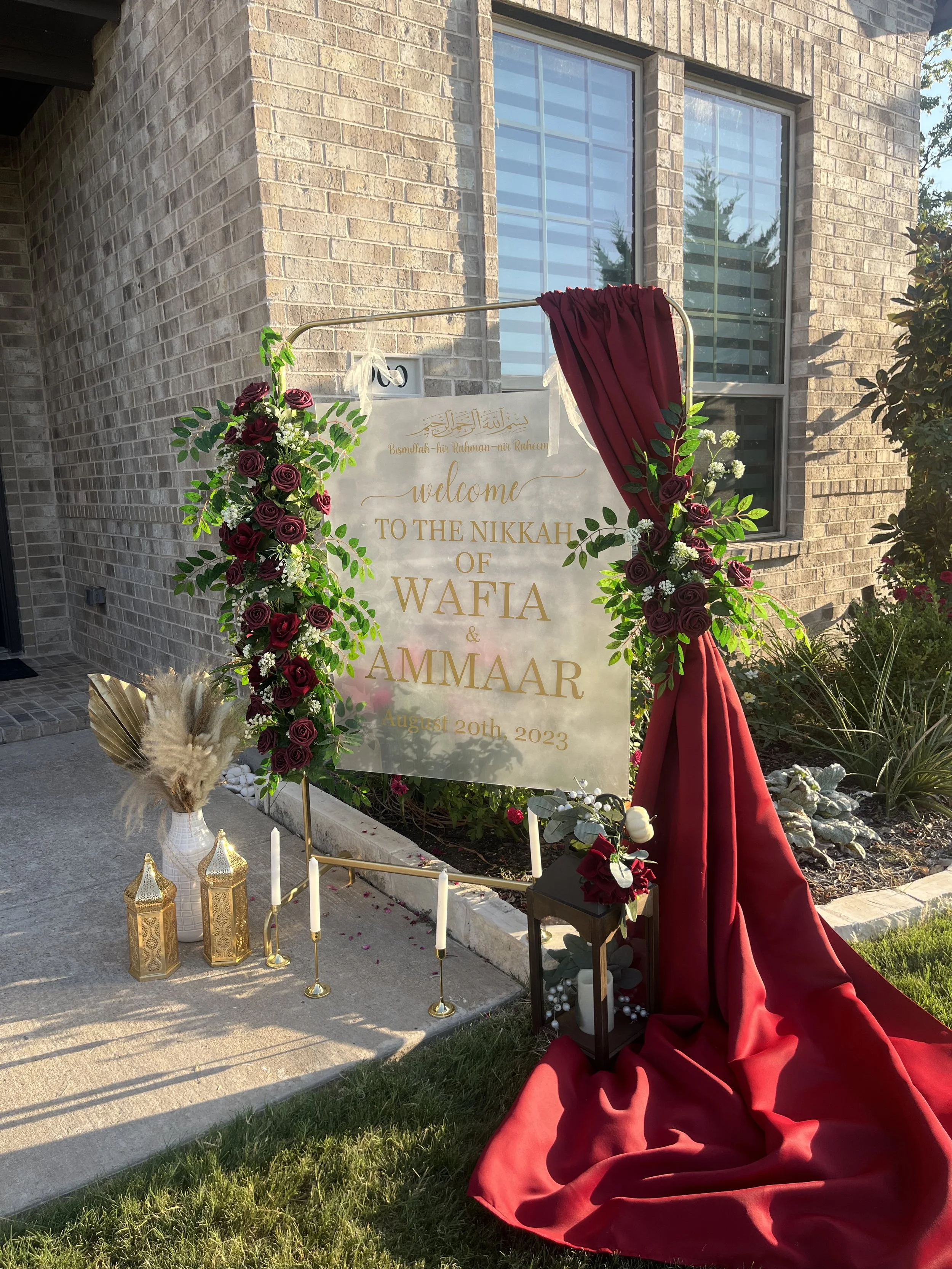 Decorative sign welcoming guests to the Nikkah of Wafia and Ammaar, dated August 20th, 2023, with floral arrangements, candles, and lanterns in an outdoor setting near a brick house.