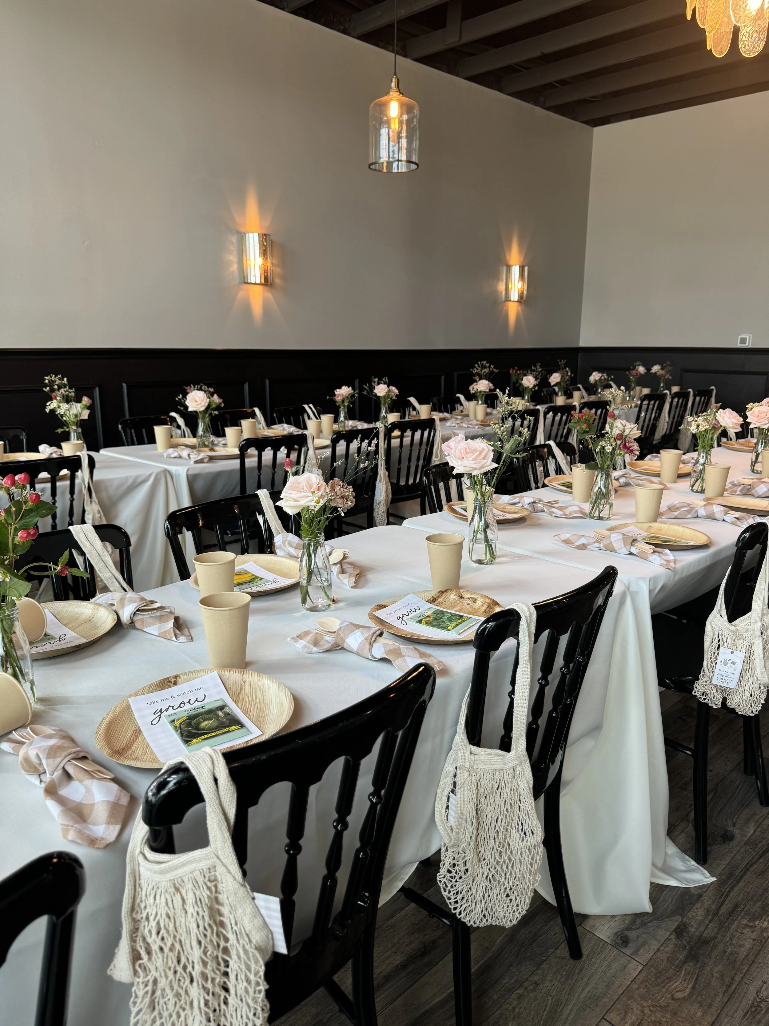 A decorated banquet-style dining setup with long tables covered in white tablecloths, adorned with small vases of pink and white flowers, beige cups, paper plates with printed napkins, and informational brochures. Black chairs with beige woven bag decorations are arranged around the tables in a room with gray walls, wood accents, and hanging pendant and wall sconces providing warm lighting.