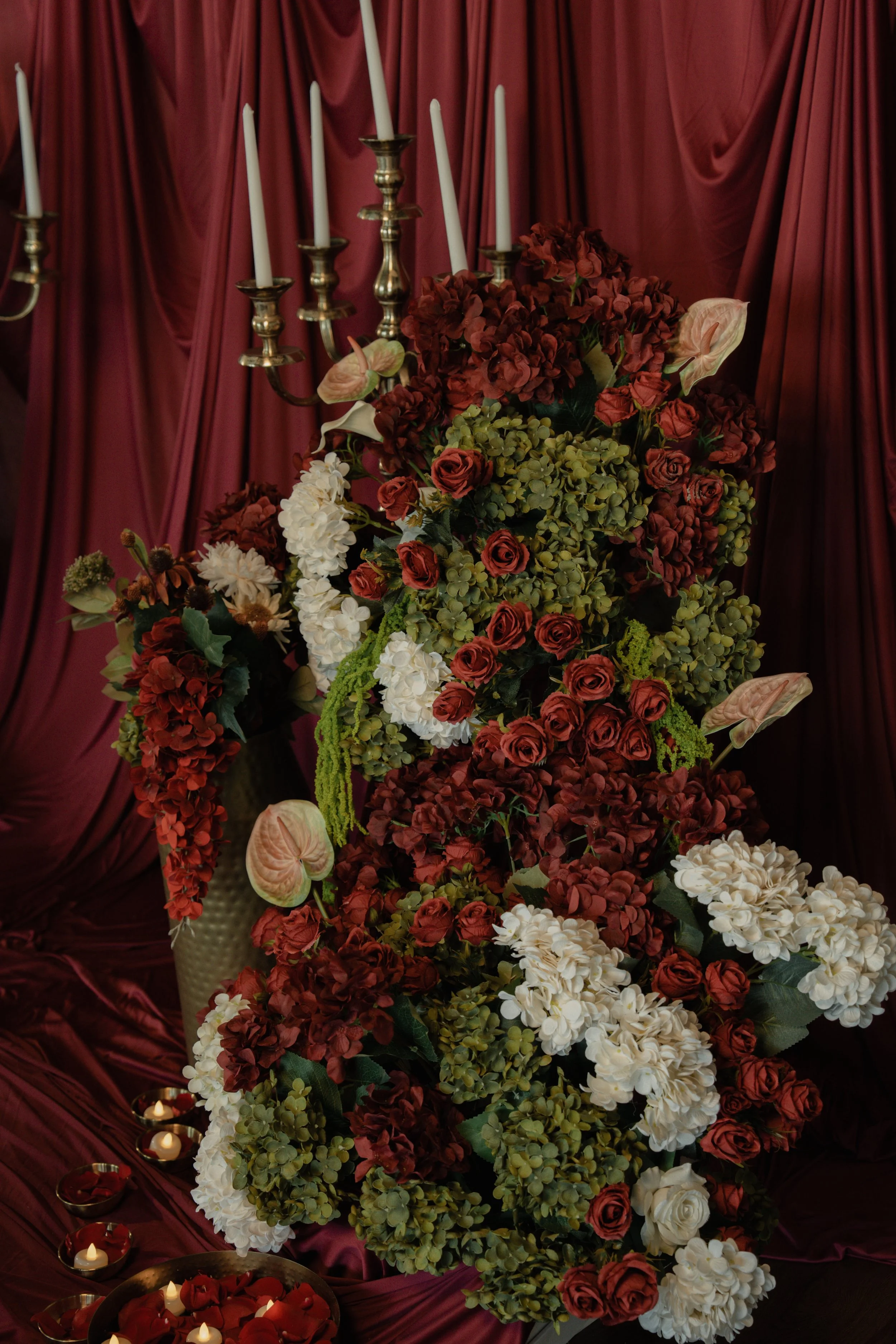A large arrangement of red, white, and green flowers, including roses and hydrangeas, with a red satin drape background and a brass candelabrum with unlit white candles. Royal wedding florals