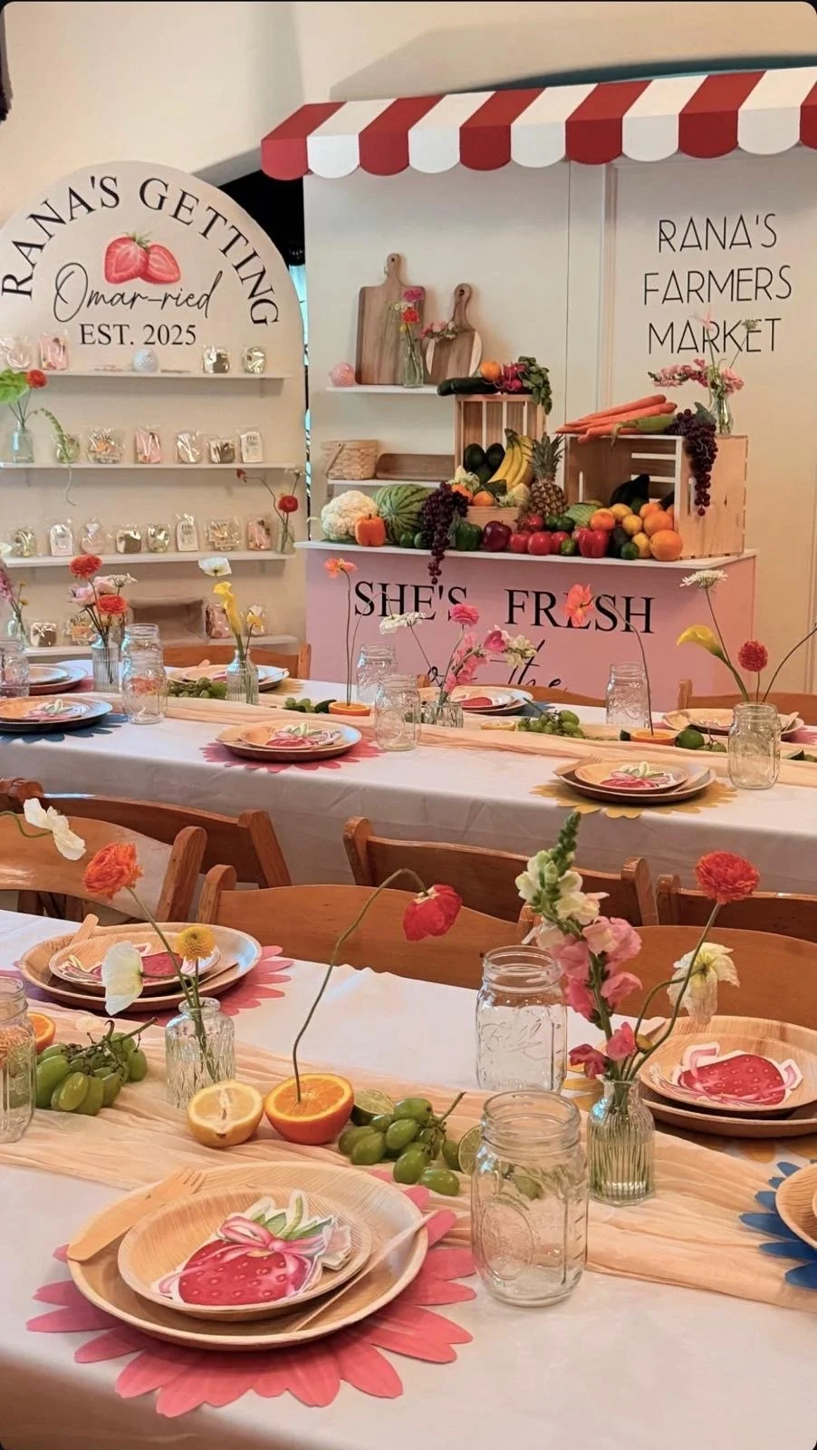 Decorative farmhouse market setup with floral centerpieces, fresh fruits, and a table set for a meal, featuring a pink sign that says "She's FRESH" and a background sign reading "Rana's Getting Mar-ried EST. 2025." The table is adorned with a pink fl