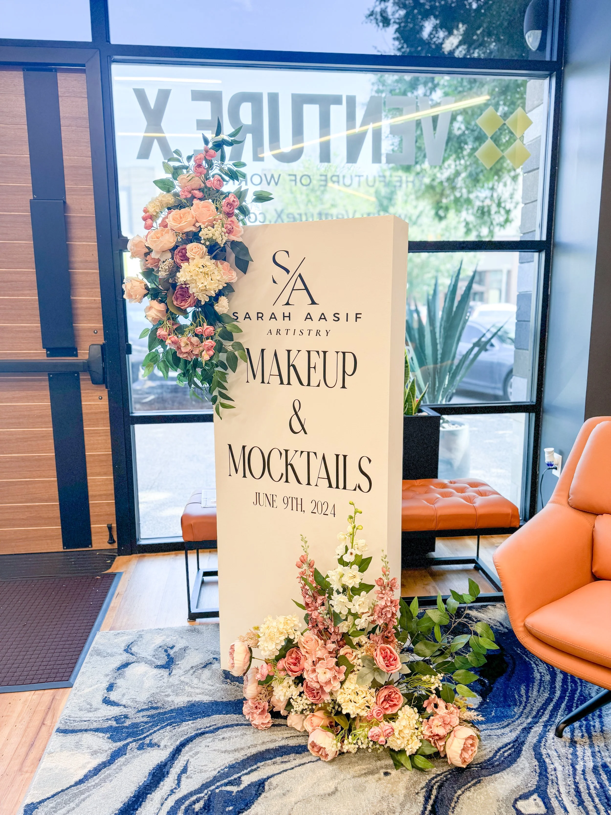 Event sign for Sarah Aasif Artistry, advertising makeup and mocktails on June 9, 2024, decorated with pink, cream, and purple flowers on and around the sign, inside a modern interior with glass windows, an orange chair, and a patterned rug.