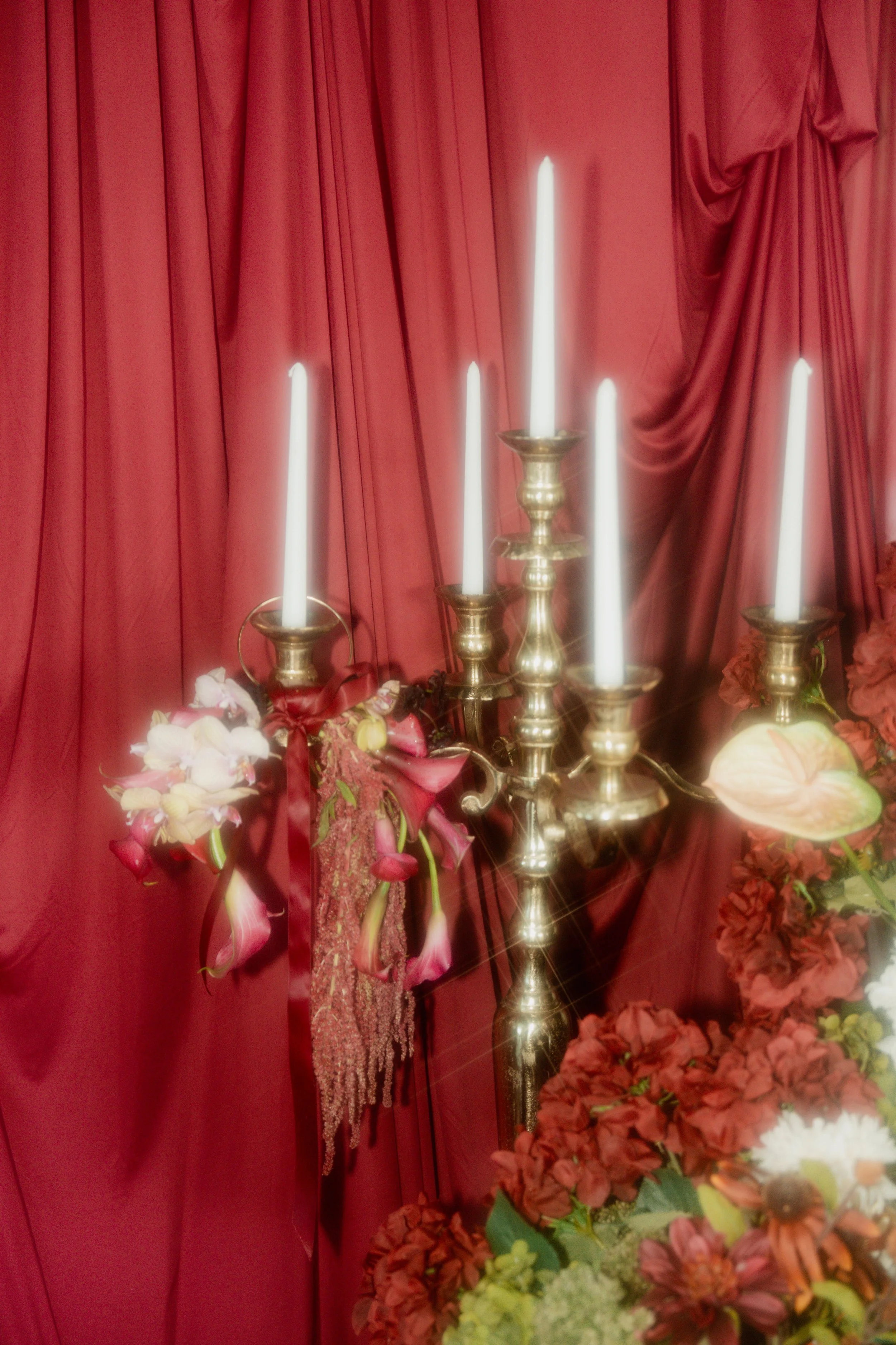 A brass candelabrum with six white candles in front of red curtains, decorated with pink and white flowers and greenery.